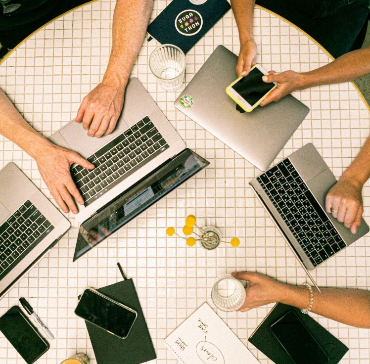 A group of people are sitting around a table with laptops and cell phones