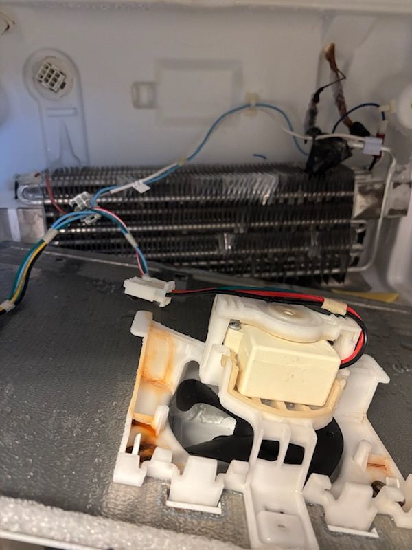 Inside of a refrigerator, exposed coils, wiring, and a fan housing with some orange staining.