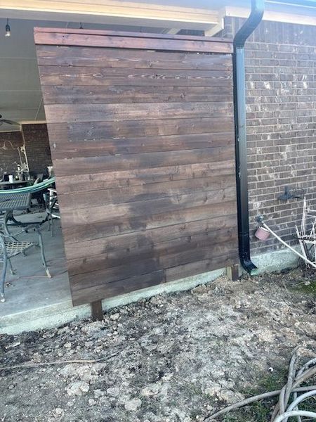 A dark stained wooden privacy screen attached to a brick building with a black gutter. The screen sits on a concrete base and a dirty yard.