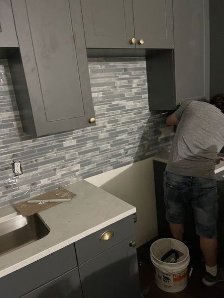 A person tiling a backsplash in a kitchen with gray cabinets, a white countertop, and gray and blue mosaic tiles.
