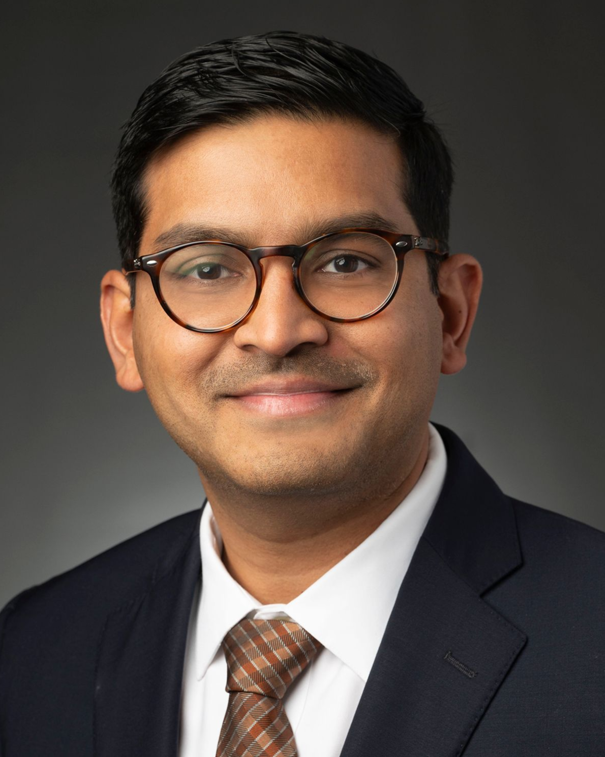 Studio headshot of a man in glasses wearing a dark suit, white shirt, and patterned tie on a gray background