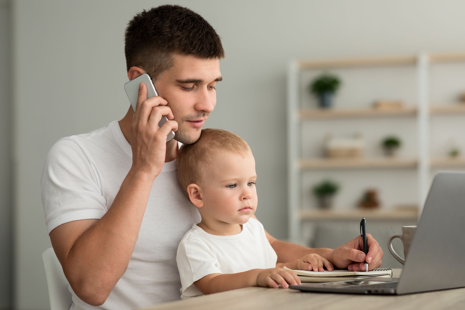 Man on phone, holding a child while working on a laptop at a desk.