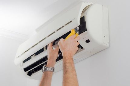 Person servicing a wall-mounted air conditioner with a screwdriver, on a white wall.