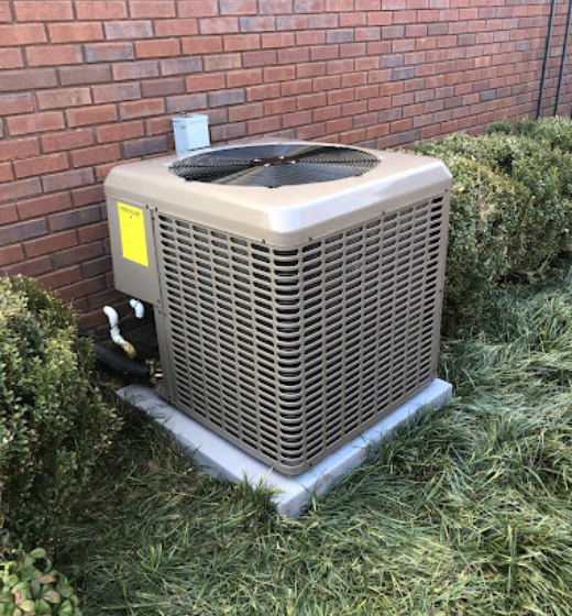 An outdoor air conditioning unit, beige, sits on a concrete pad by a brick wall and greenery.