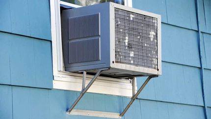Gray air conditioner unit in a window, supported by metal brackets, on a blue house.