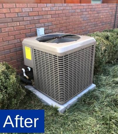 An air conditioning unit, beige and boxy, sits on a concrete pad next to a brick wall and green bushes.