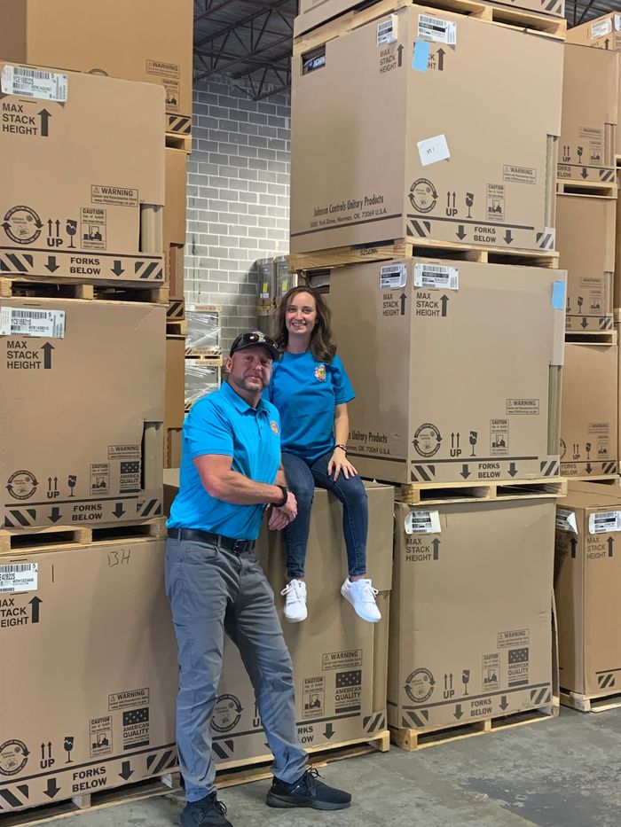 Man and woman in warehouse pose near stacked cardboard boxes. Man smiles, woman sits on a box.