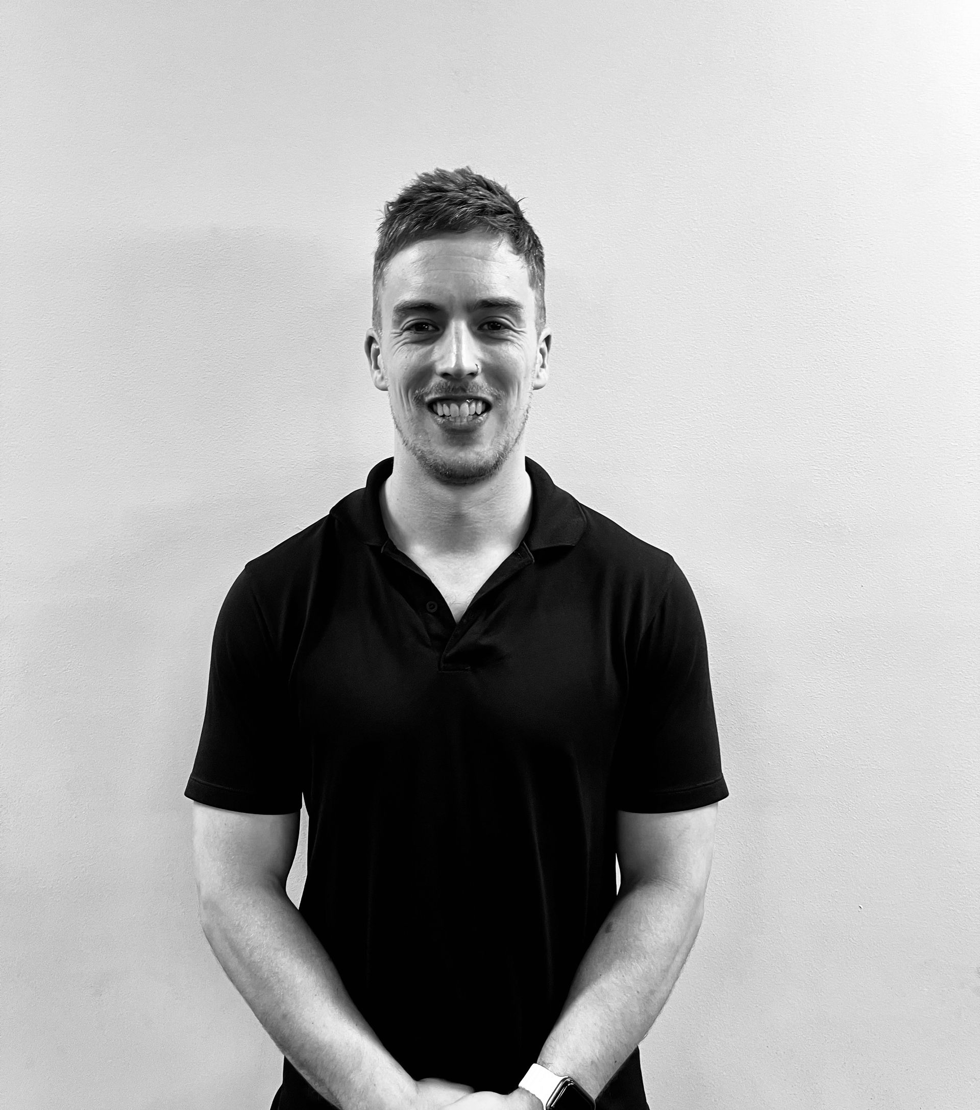 Man in black polo shirt smiles, arms crossed, standing in front of a light wall.