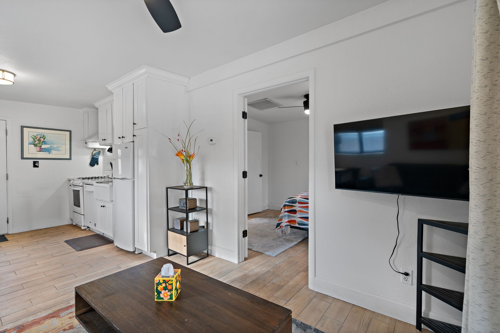Living room with TV on wall, open doorway, and kitchen area partially visible.