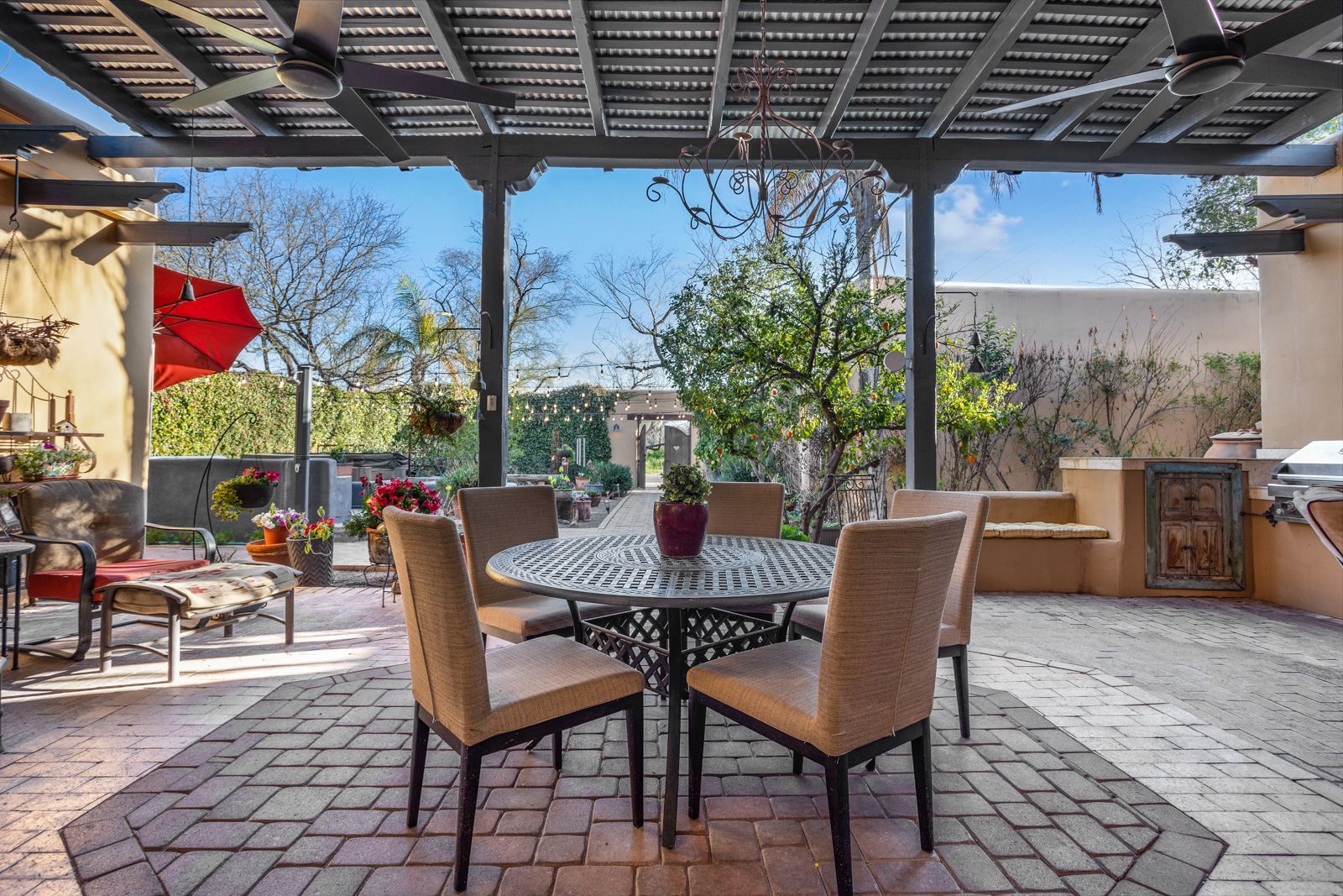 Patio with brick flooring, a table, and chairs under a shaded structure, with outdoor decor and greenery visible.