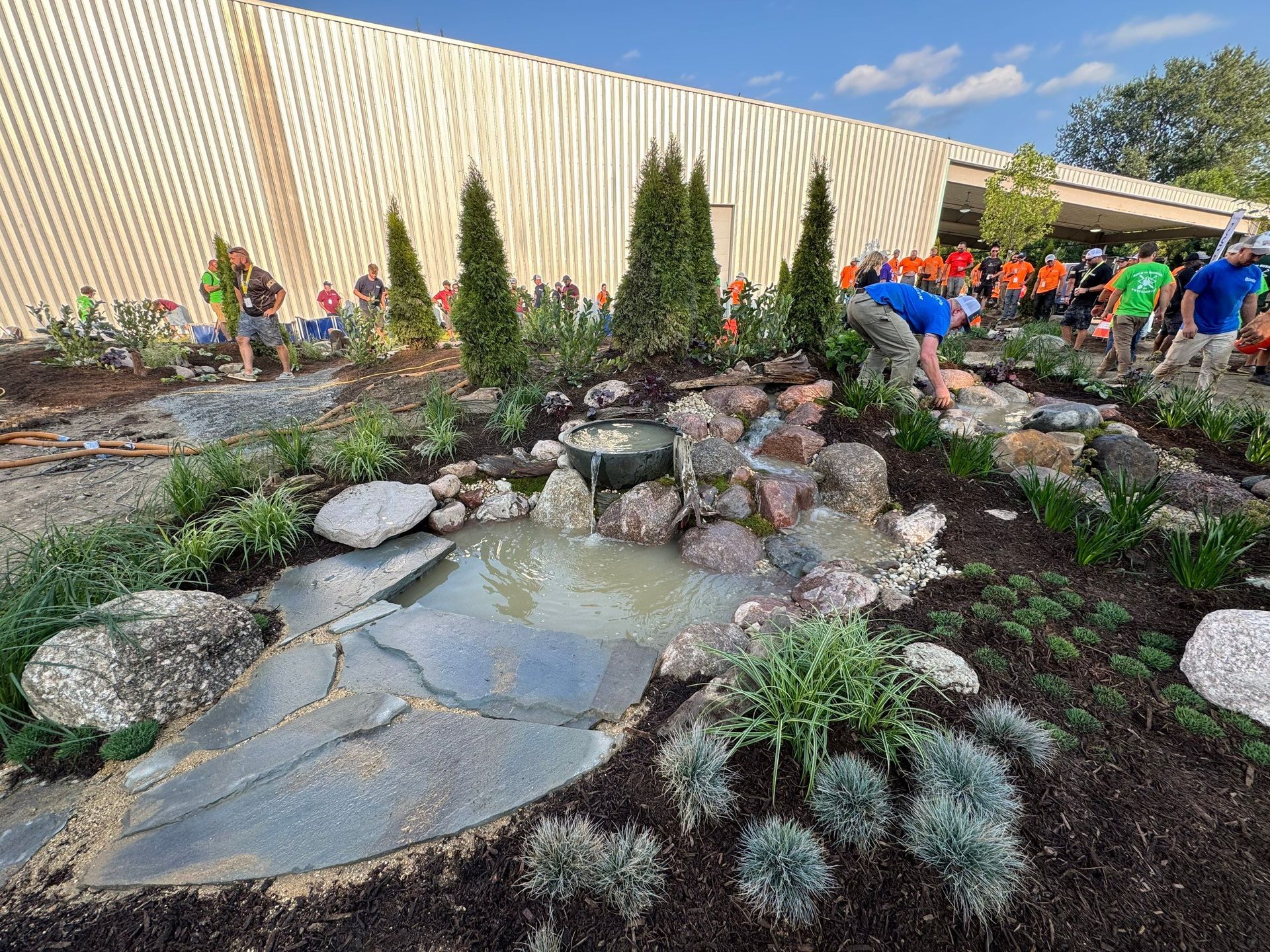 People in bright shirts work on a garden with a small waterfall, surrounded by rocks and lush greenery, near a beige industrial building.