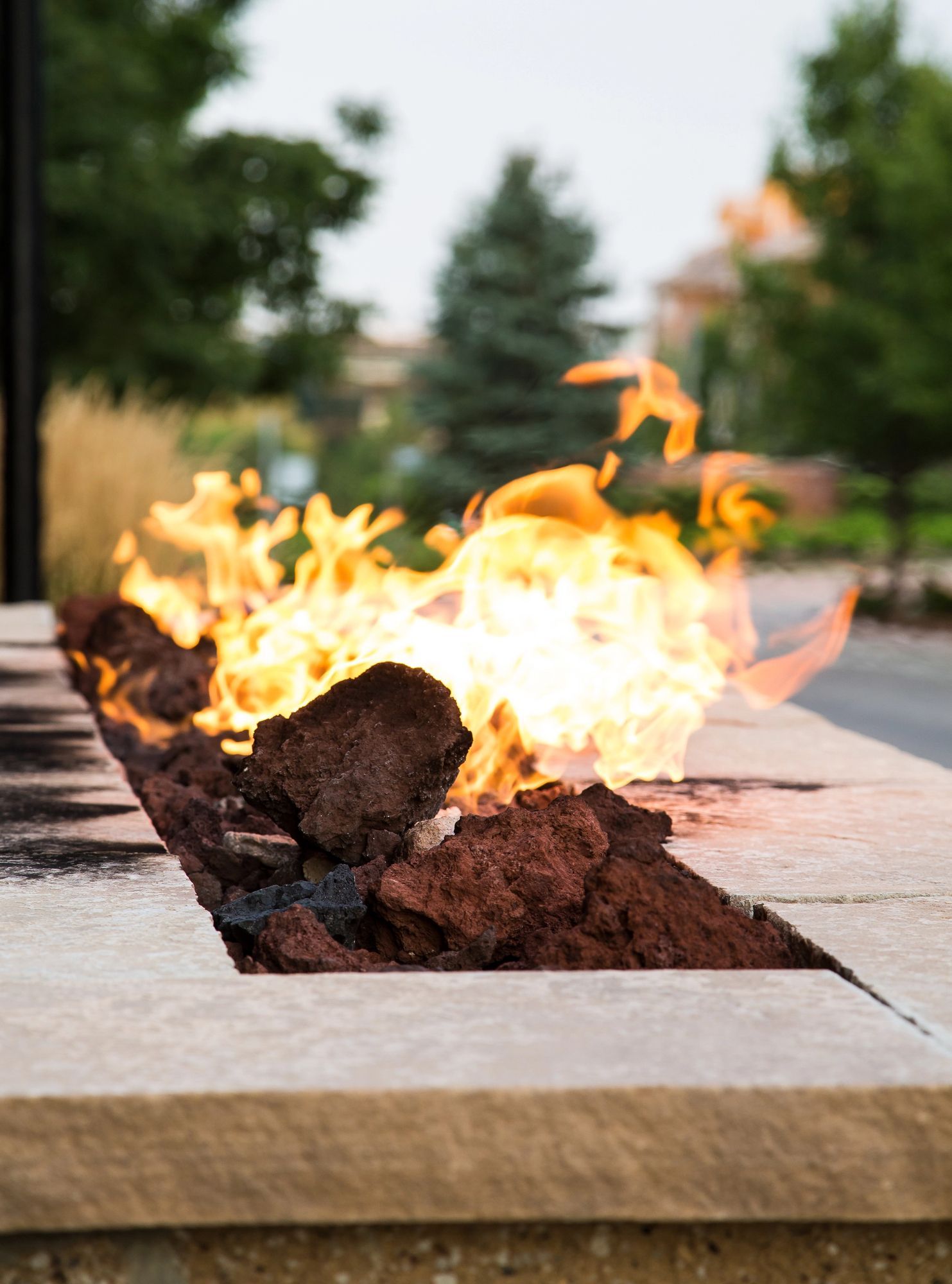 Flames erupt from a stone fire pit, with a blurred background of trees and buildings.