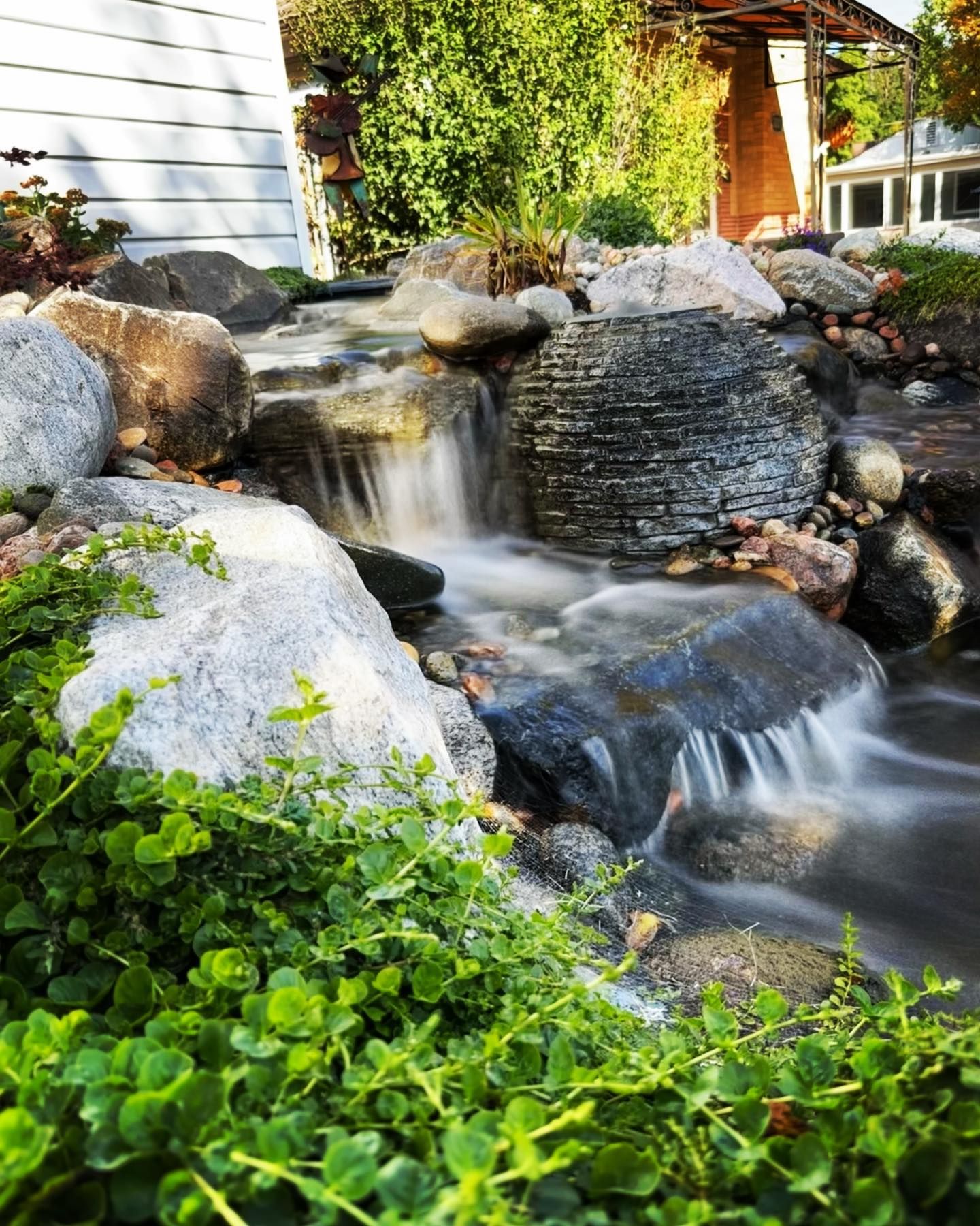Waterfall cascading over rocks in a lush garden, with green foliage and a glimpse of a house in the background.