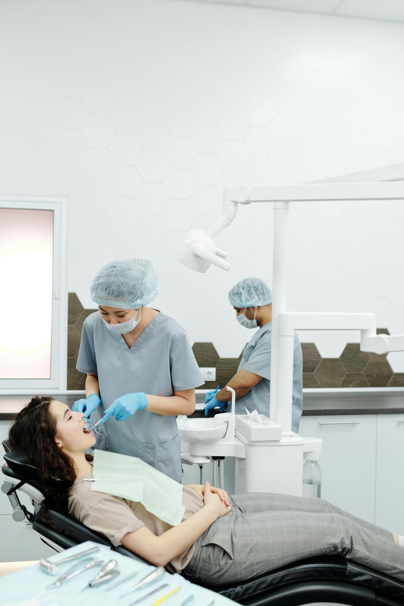 Patient in dentist chair, mouth open; dentist examining teeth with instruments in bright, sterile room.