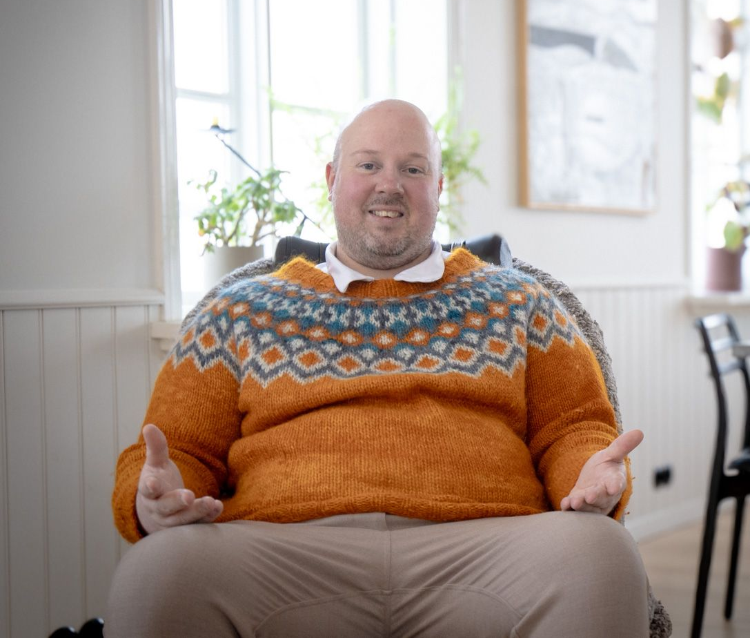 A man is sitting in a chair talking to another man.