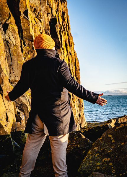 A man in a black coat and yellow hat is standing on a rocky cliff overlooking the ocean.