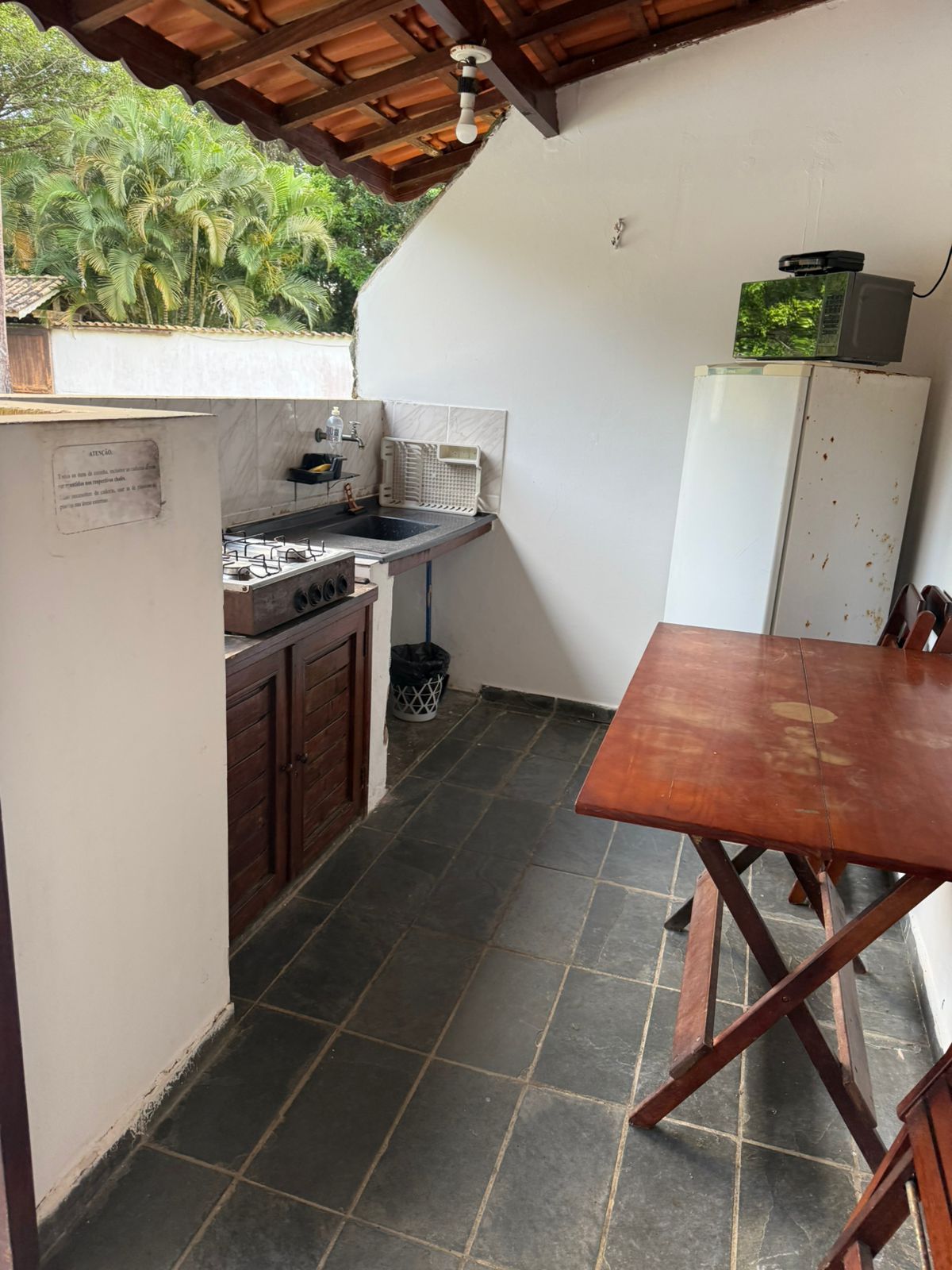 Cozinha externa com fogão, pia, geladeira e mesa. Piso de cerâmica escura, paredes brancas e armários de madeira.