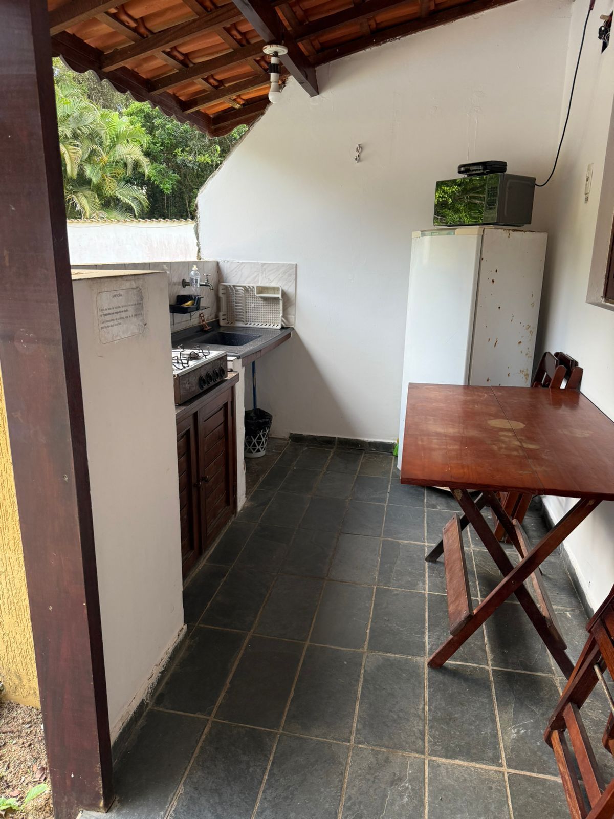 Cozinha externa com fogão, pia, geladeira, mesa e piso de cerâmica escura.