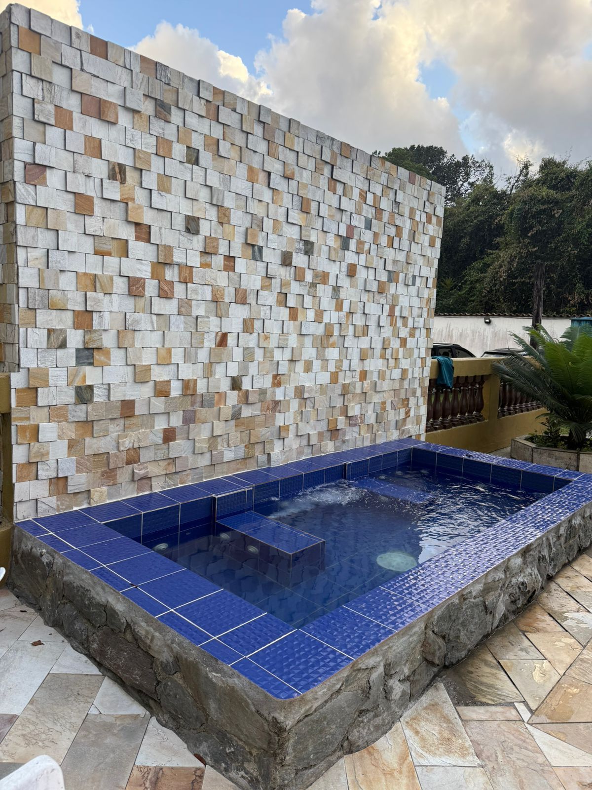 Pequena piscina com azulejos azuis e jatos de hidromassagem em frente a uma parede de pedra texturizada; ao ar livre.