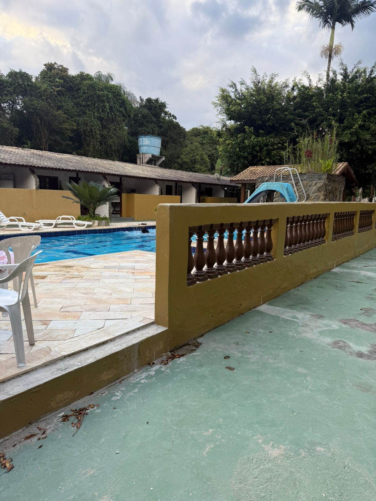Piscina ao lado de um muro amarelo com grades decorativas, prédios e árvores. Céu nublado.