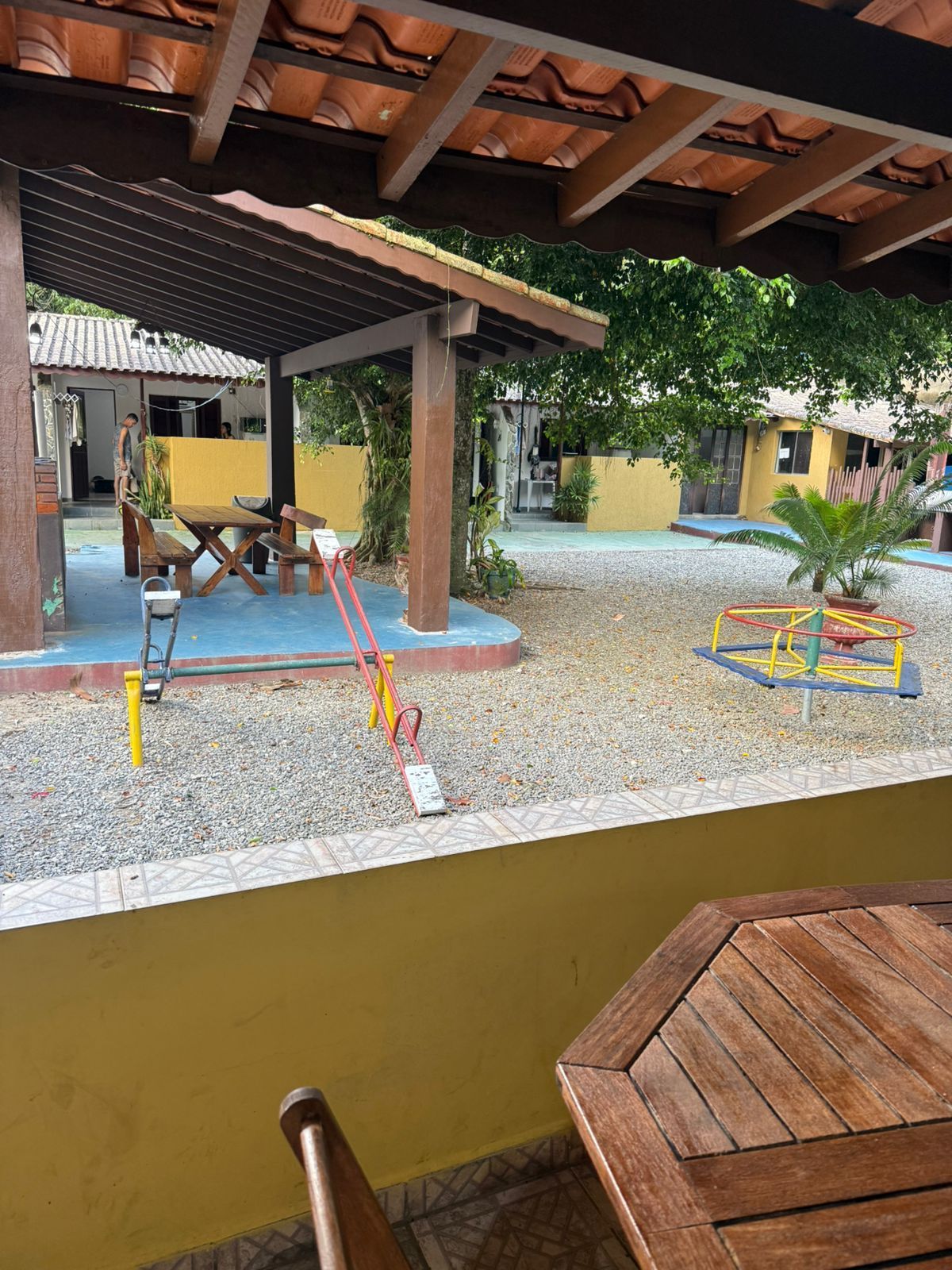 Parque infantil com balanço e carrossel, visto de um pátio coberto com mesa de madeira.