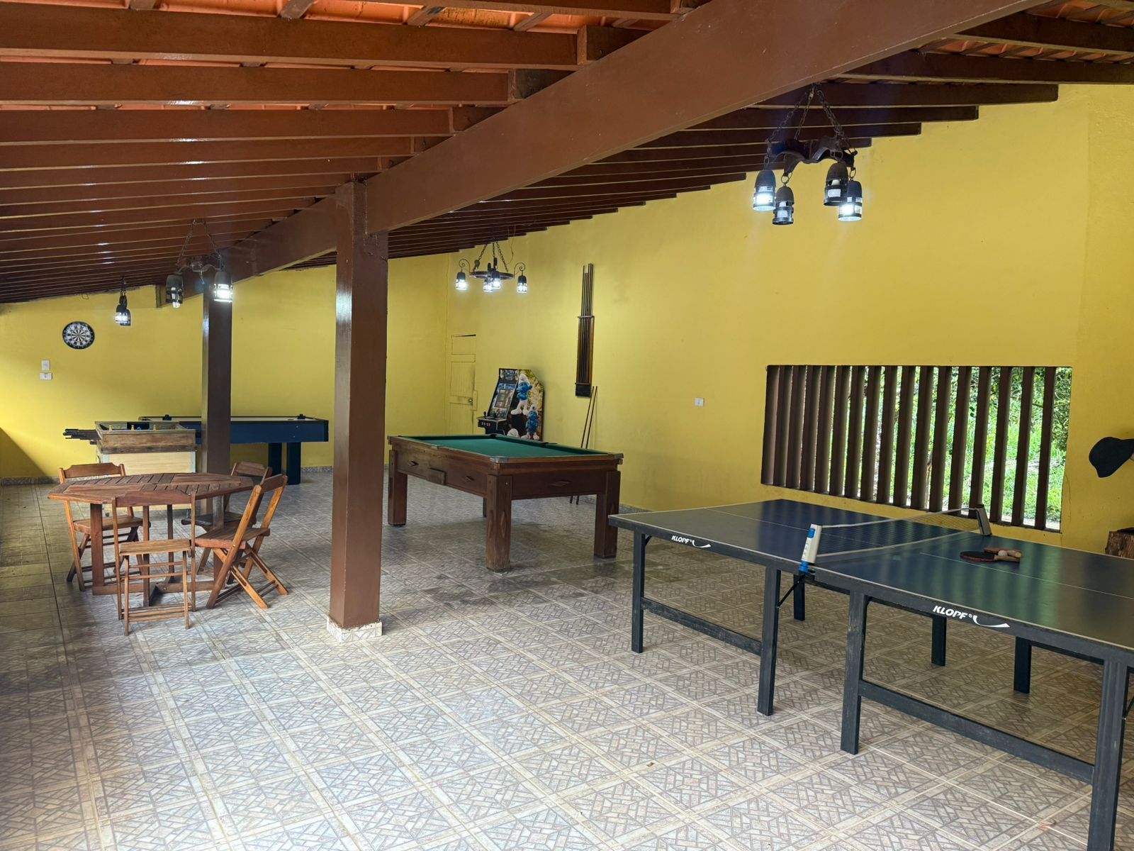 Sala de jogos com paredes amarelas, piso de cerâmica e diversas mesas de jogos, incluindo sinuca e pingue-pongue.