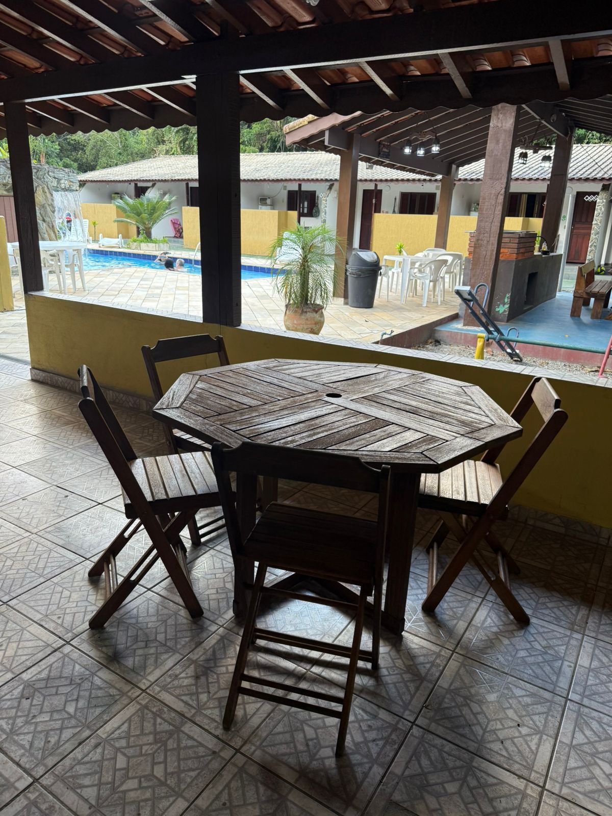 Mesa e cadeiras de madeira em um pátio de azulejos, com vista para uma piscina e um prédio.