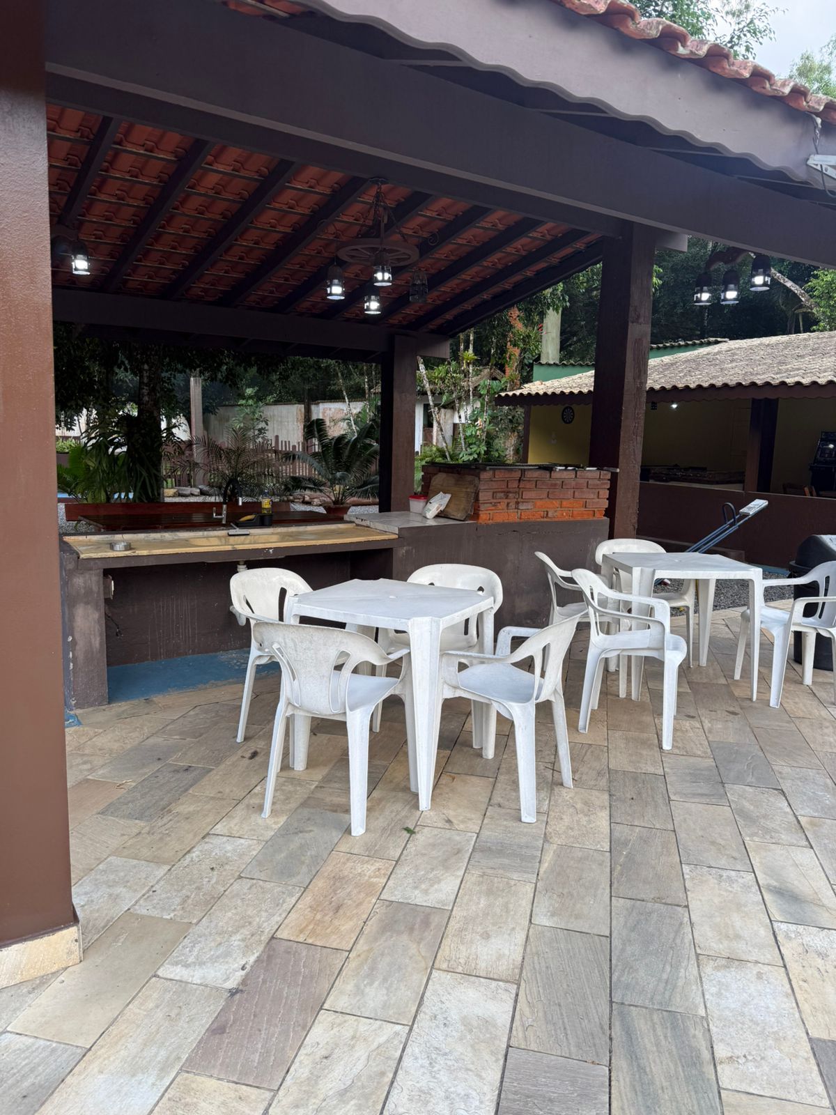 Área de pátio com mesas brancas, cadeiras e uma churrasqueira coberta sob um telhado marrom.