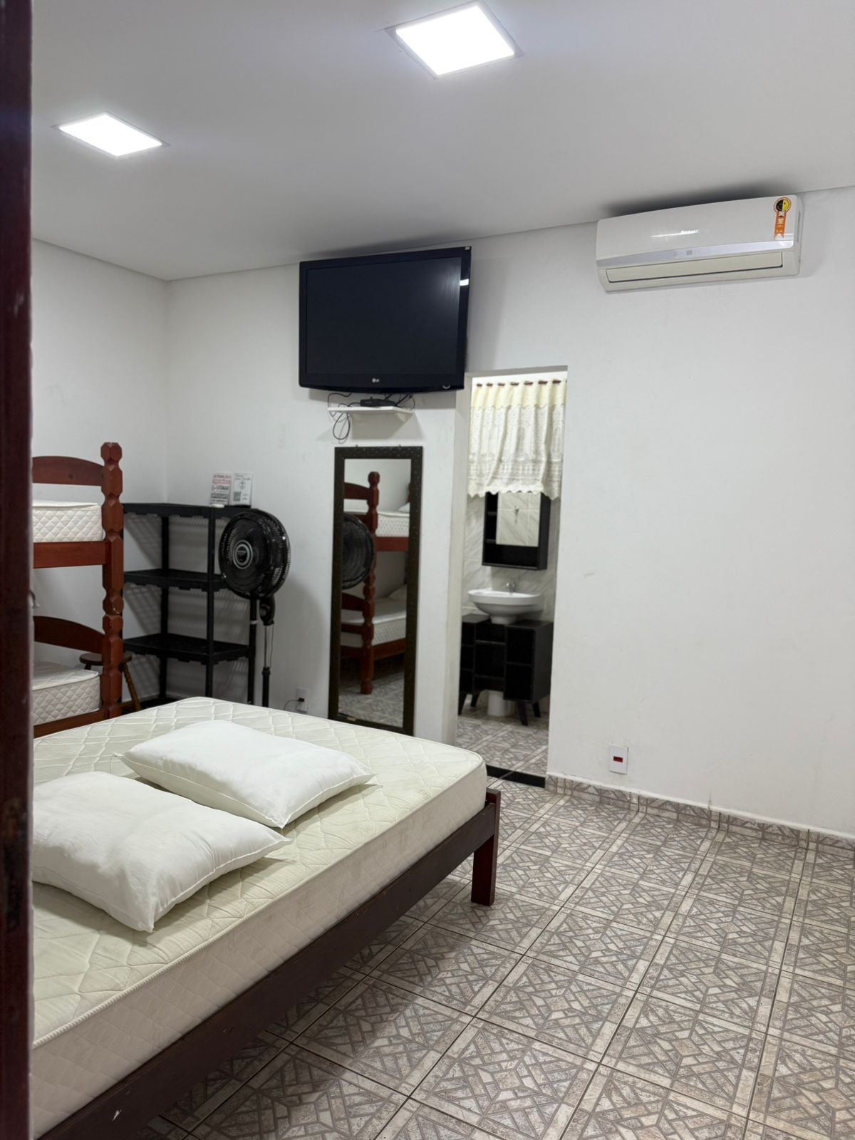 Quarto com cama, TV, beliches, espelho e ar condicionado. Paredes brancas, piso estampado.