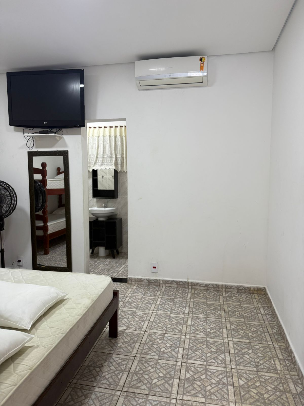 Quarto com cama, TV, ar-condicionado, espelho com beliches e banheiro. Piso de ladrilho com padrão bege.