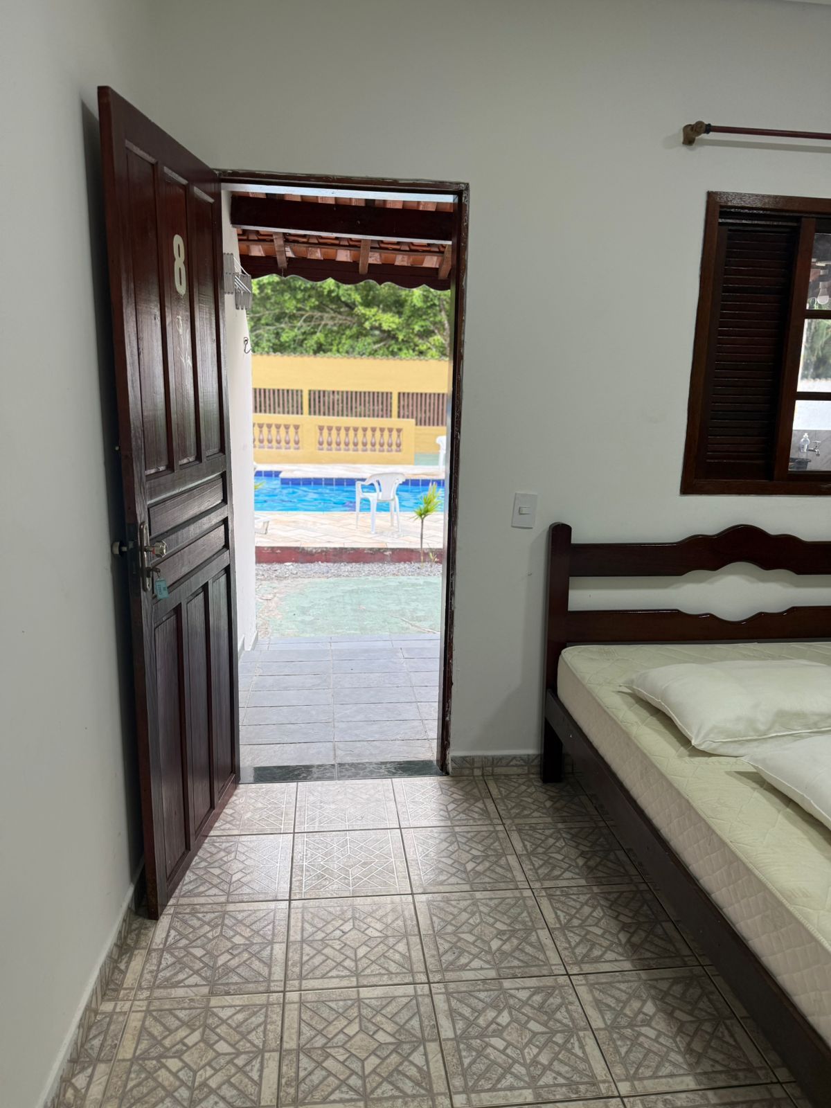 A porta se abre para uma piscina. Lá dentro, uma cama, piso de cerâmica e uma janela com venezianas fechadas.