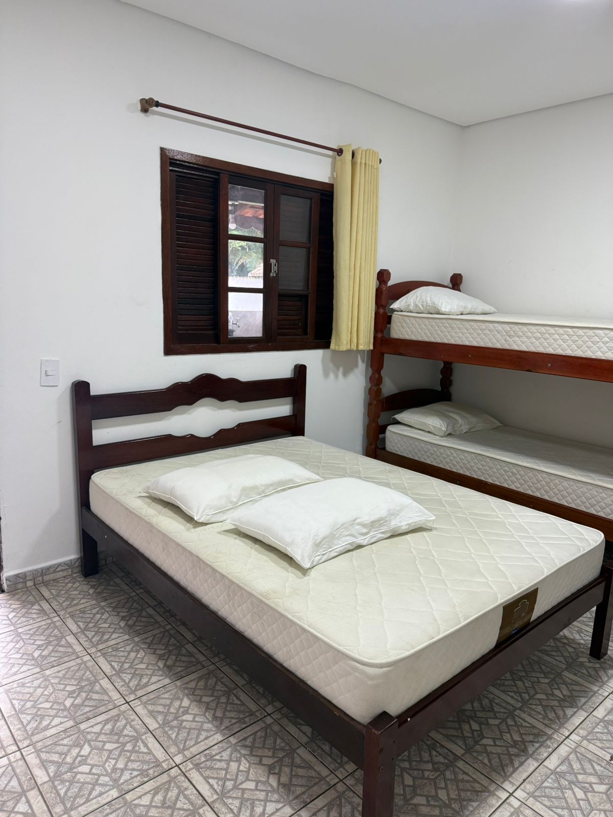 Quarto com cama de casal, beliches, janela com persianas de madeira e piso de cerâmica estampada.