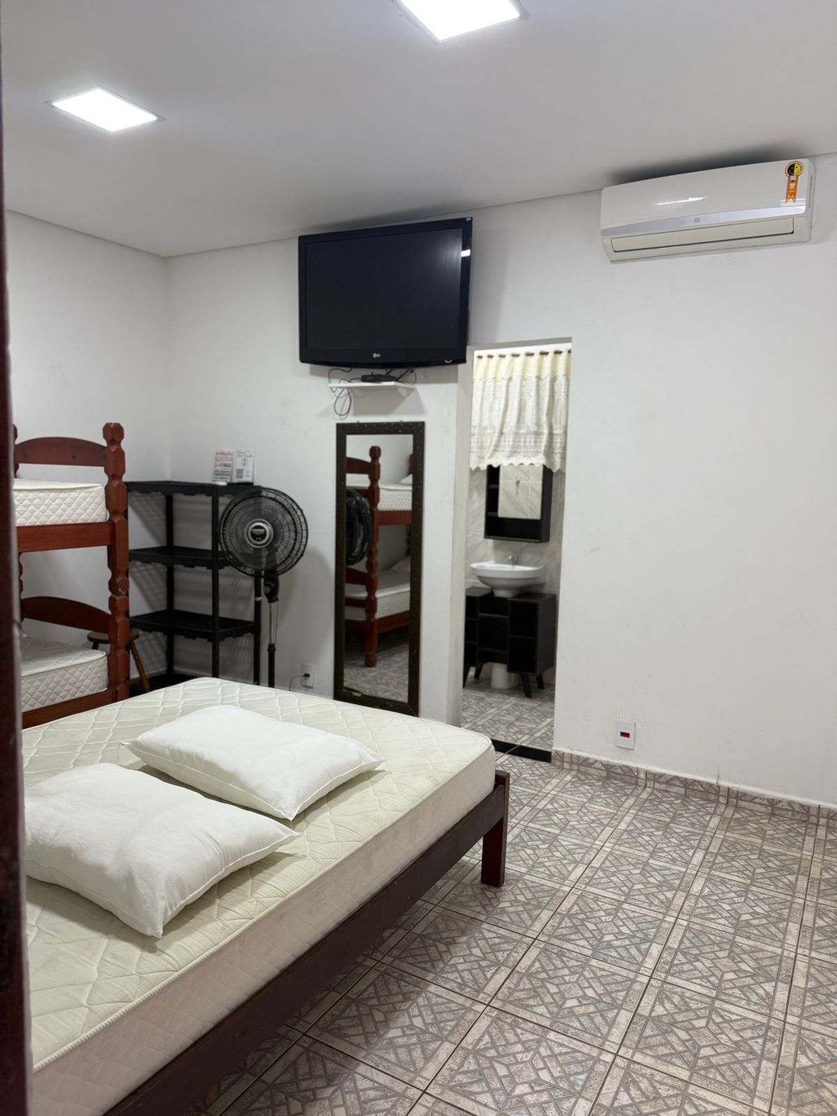 Quarto com cama, beliches, TV e espelho refletindo o banheiro.