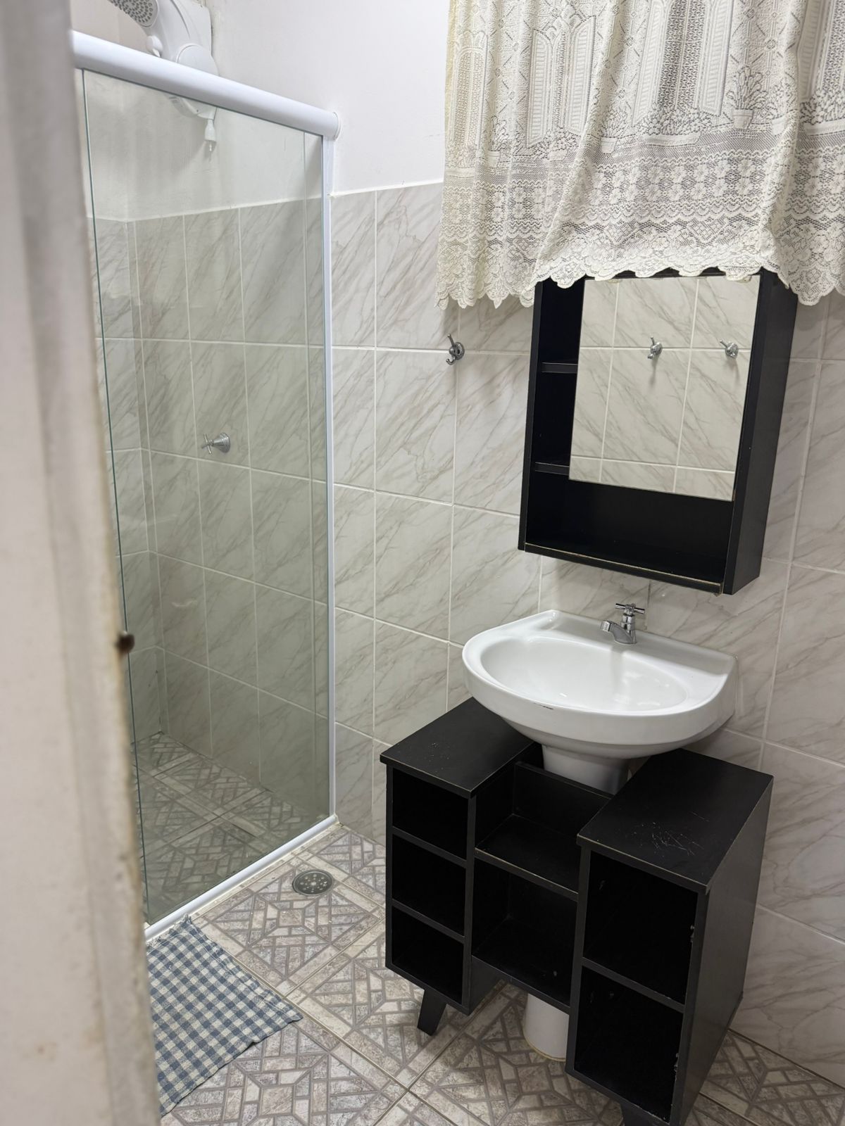 Banheiro pequeno com chuveiro, pia e armário. Paredes e piso de azulejos cinza e branco.