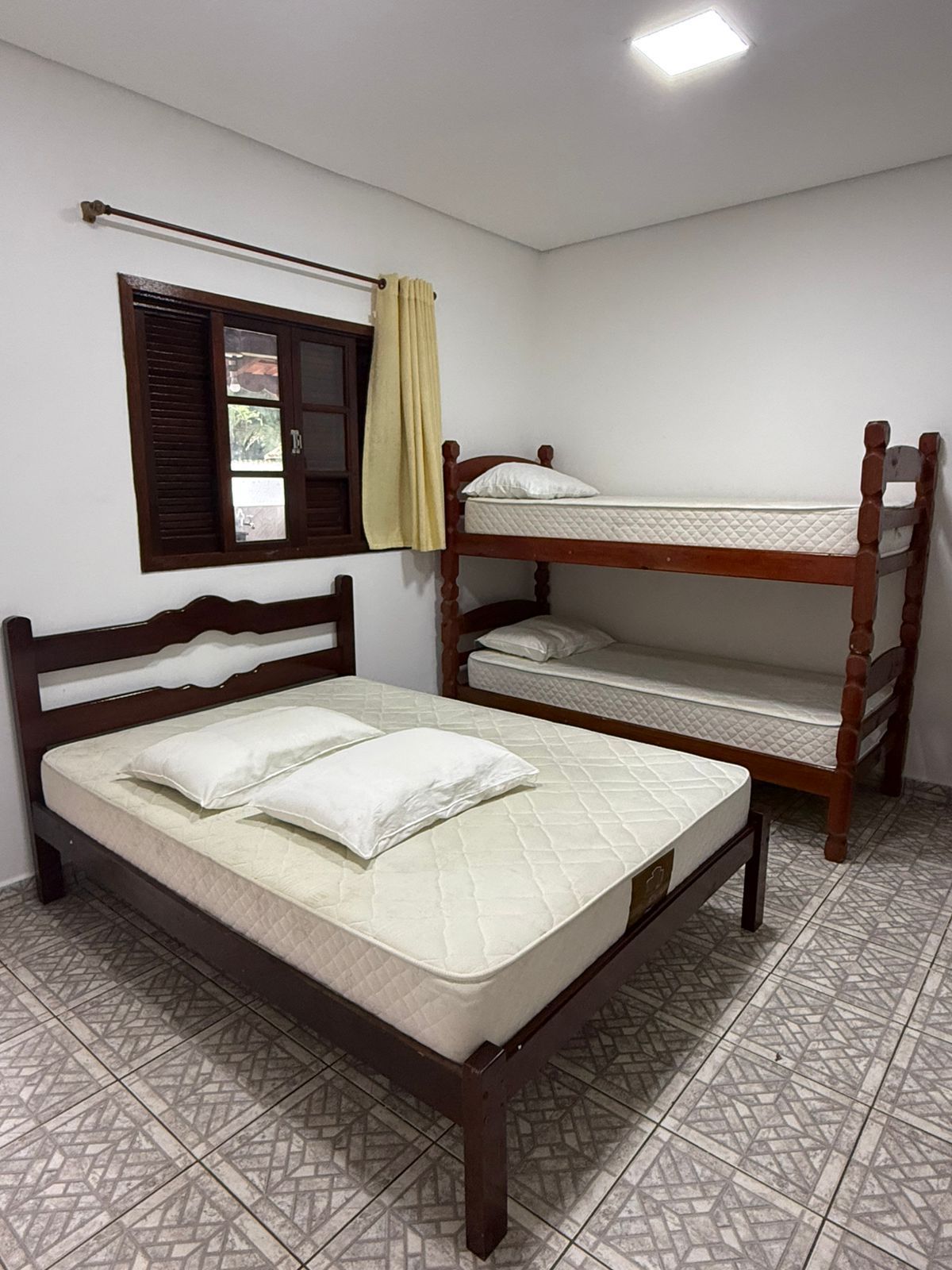 Quarto com uma cama de casal e um beliche. Paredes brancas, piso de cerâmica e uma janela.