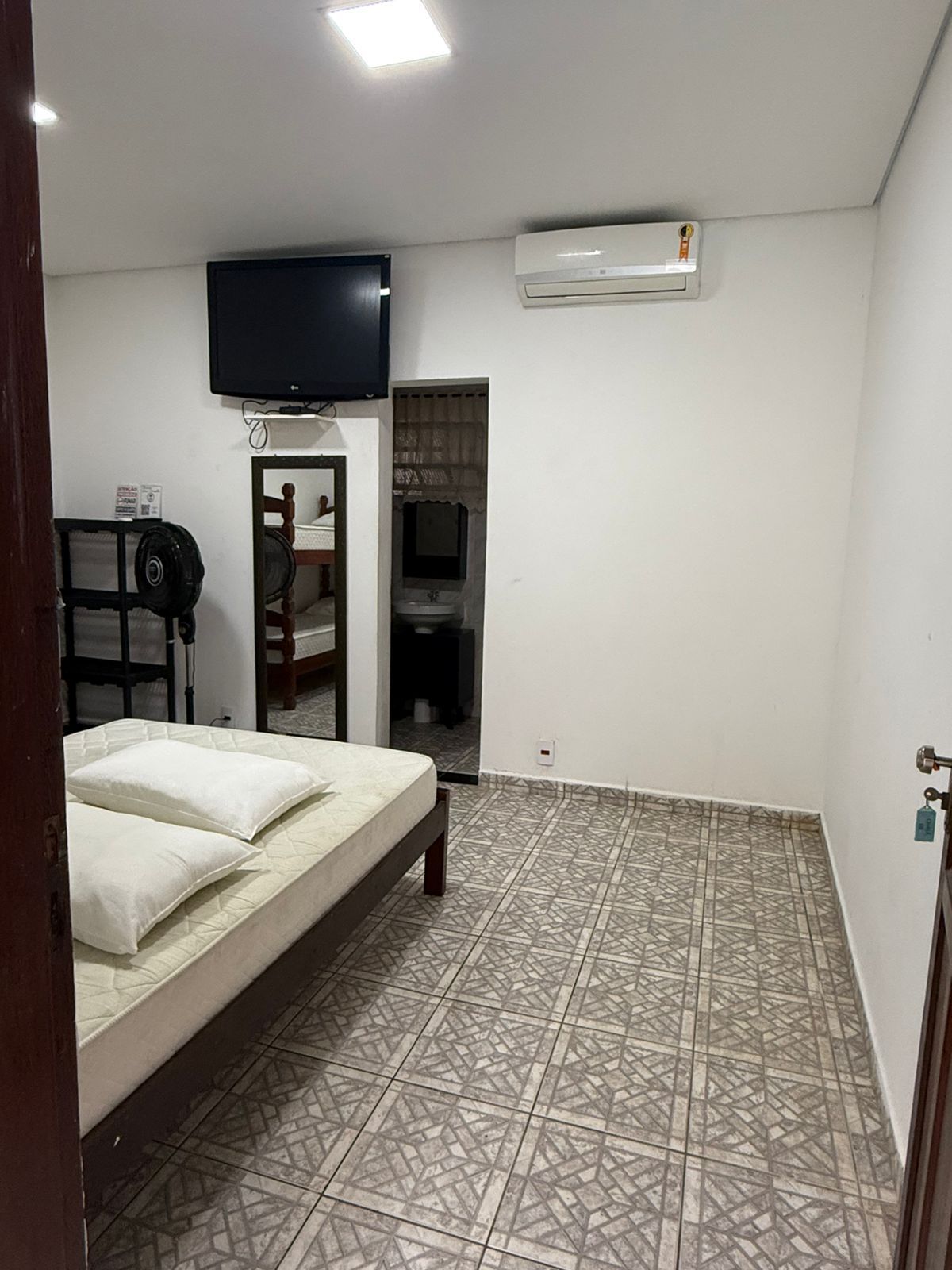 Quarto com cama, TV e banheiro visíveis. Piso cinza estampado. Paredes brancas.