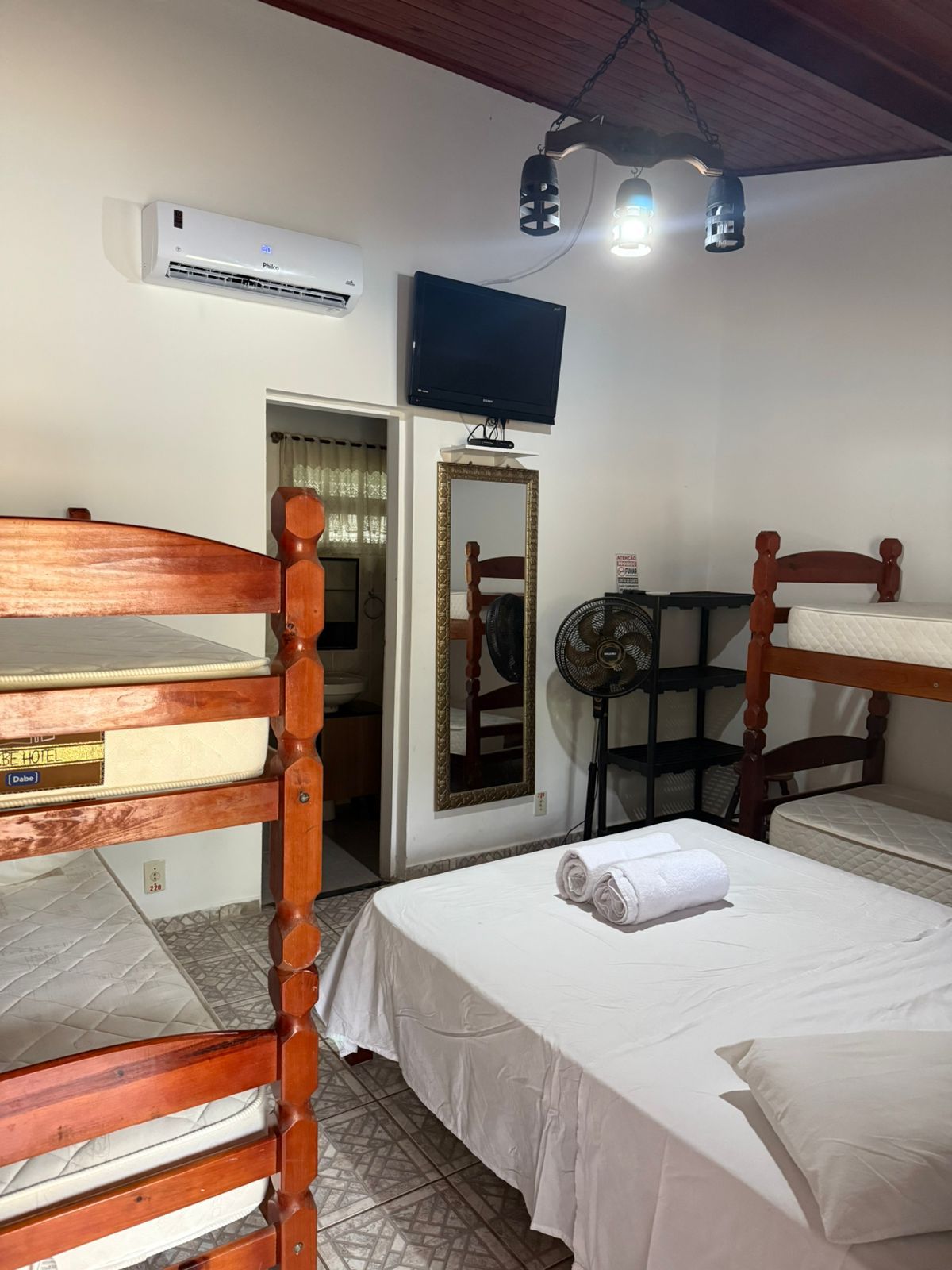 Quarto com beliches, uma cama de casal, TV, espelho, ventilador e ar-condicionado. Paredes e roupa de cama brancas.