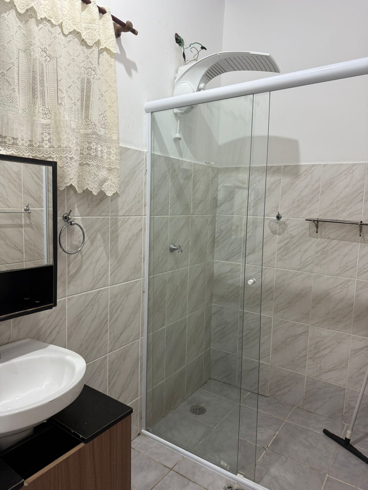 Banheiro pequeno com chuveiro, pia e espelho. Azulejos brancos e bege, box de vidro.