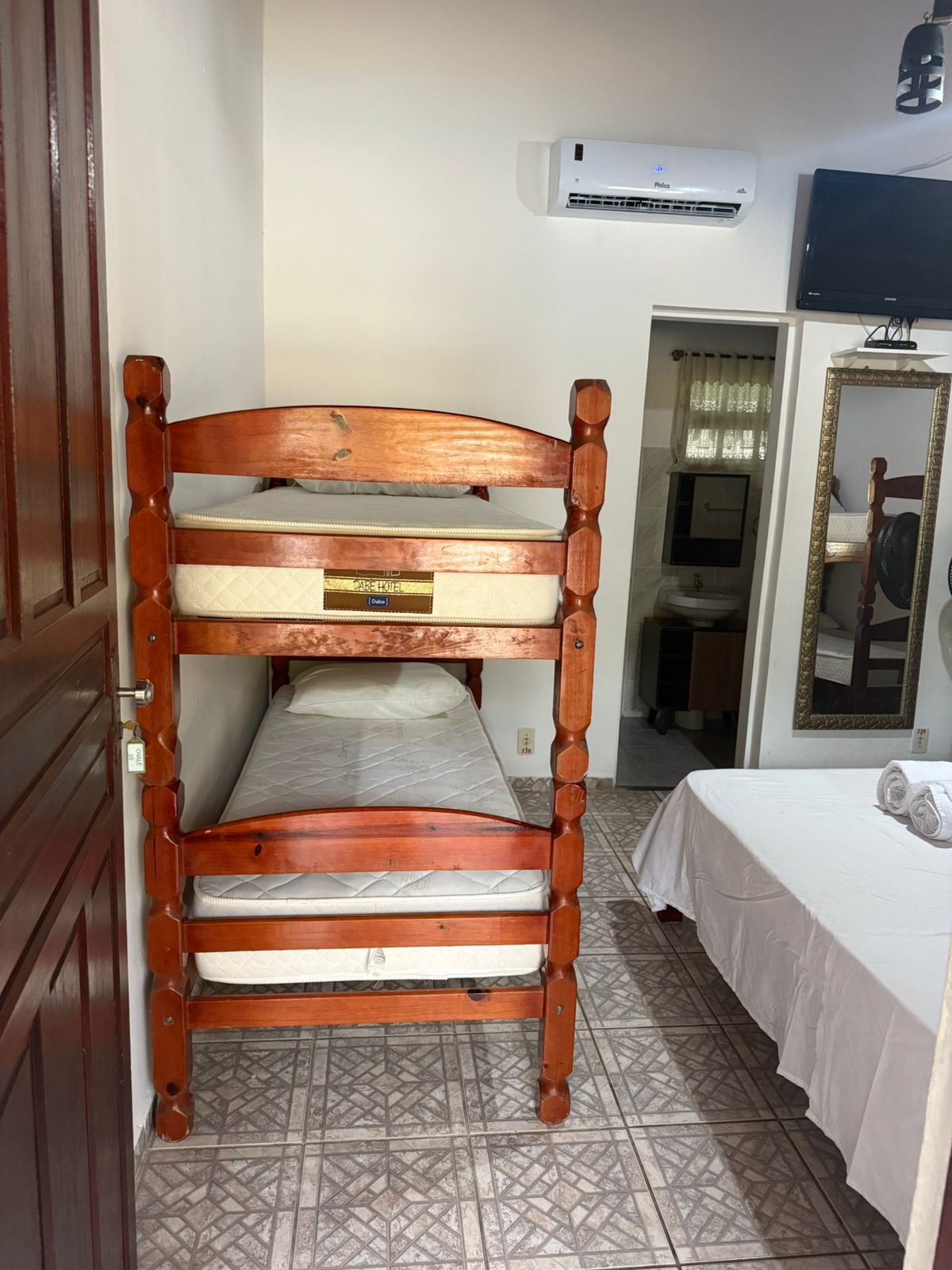 Beliches em um quarto com cama branca, espelho, TV e banheiro aberto.