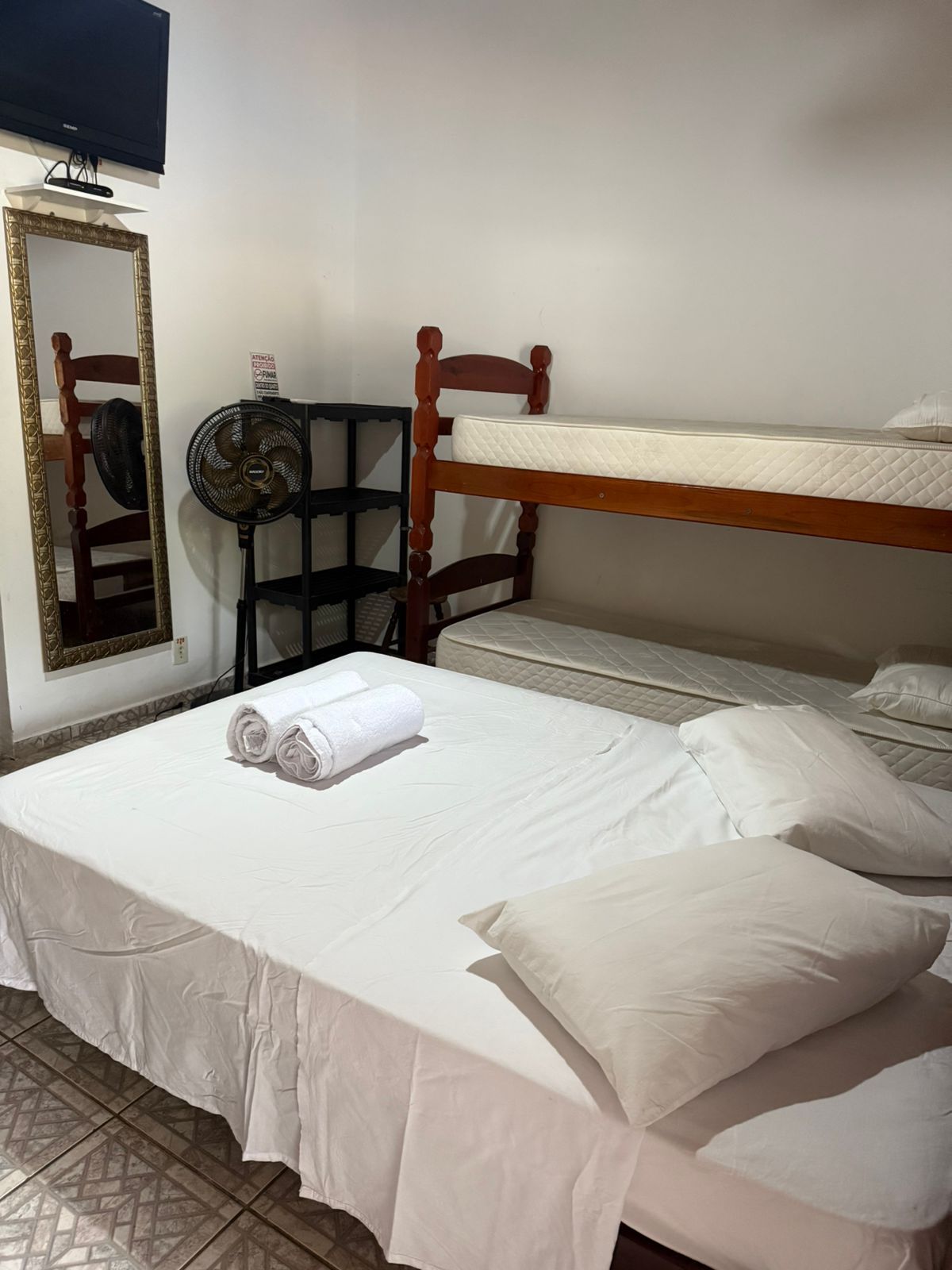 Quarto com beliches, cama de casal com lençóis brancos e espelho alto.