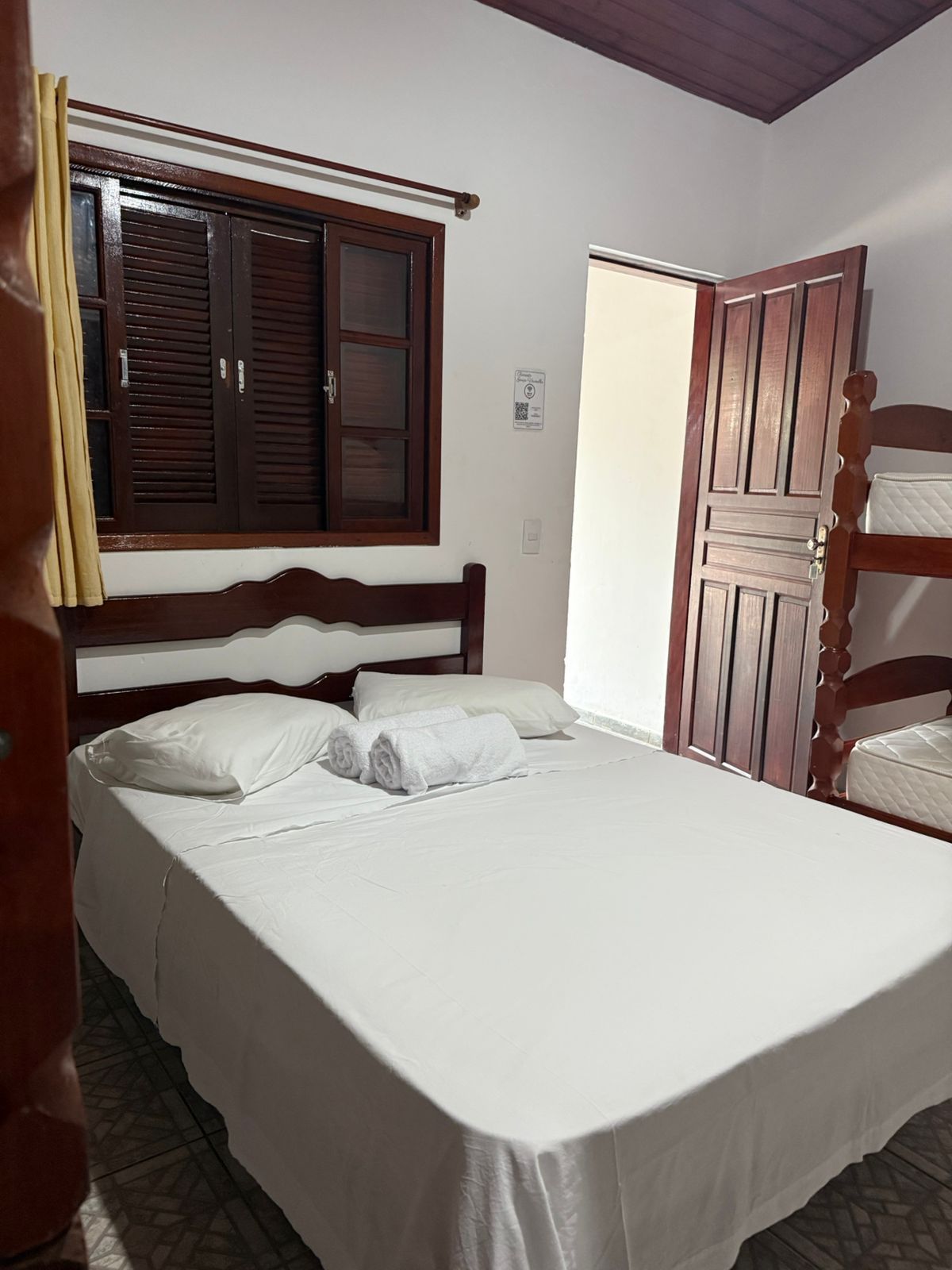 Quarto com cama de casal e beliche, janela, porta aberta. Roupa de cama branca, móveis de madeira escura.