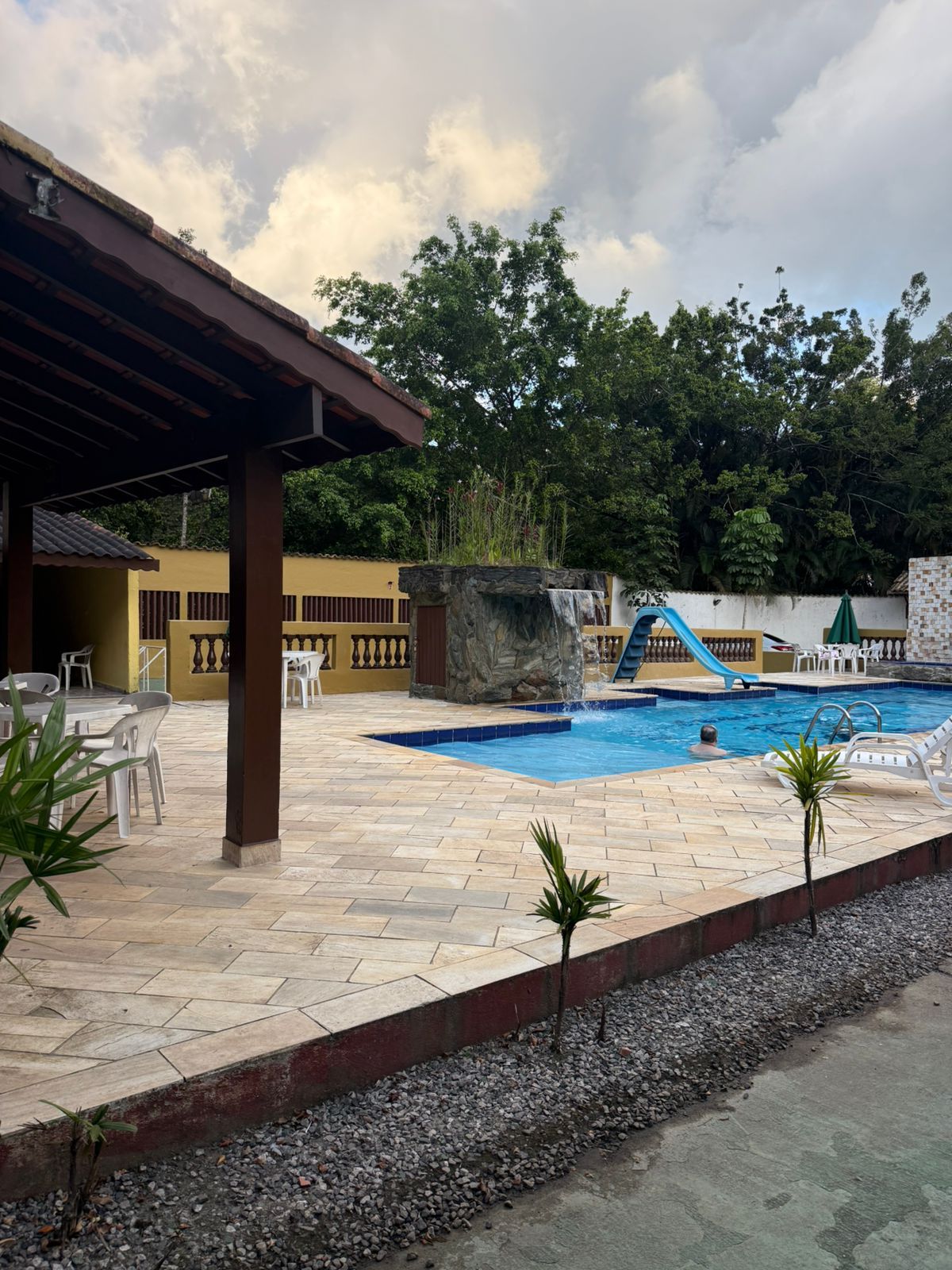 Área de piscina com água azul, árvores e um pequeno escorregador sob um céu nublado.