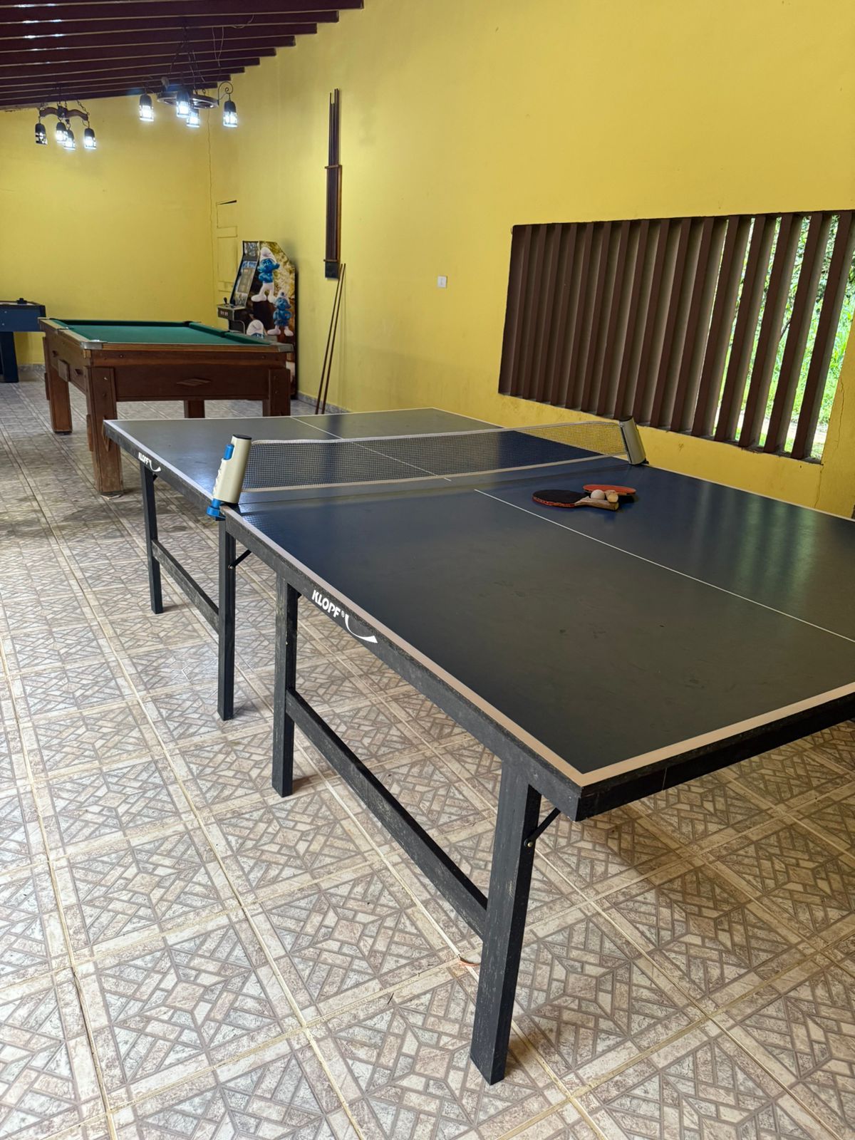 Mesa de pingue-pongue em uma sala amarela com uma mesa de sinuca ao fundo.