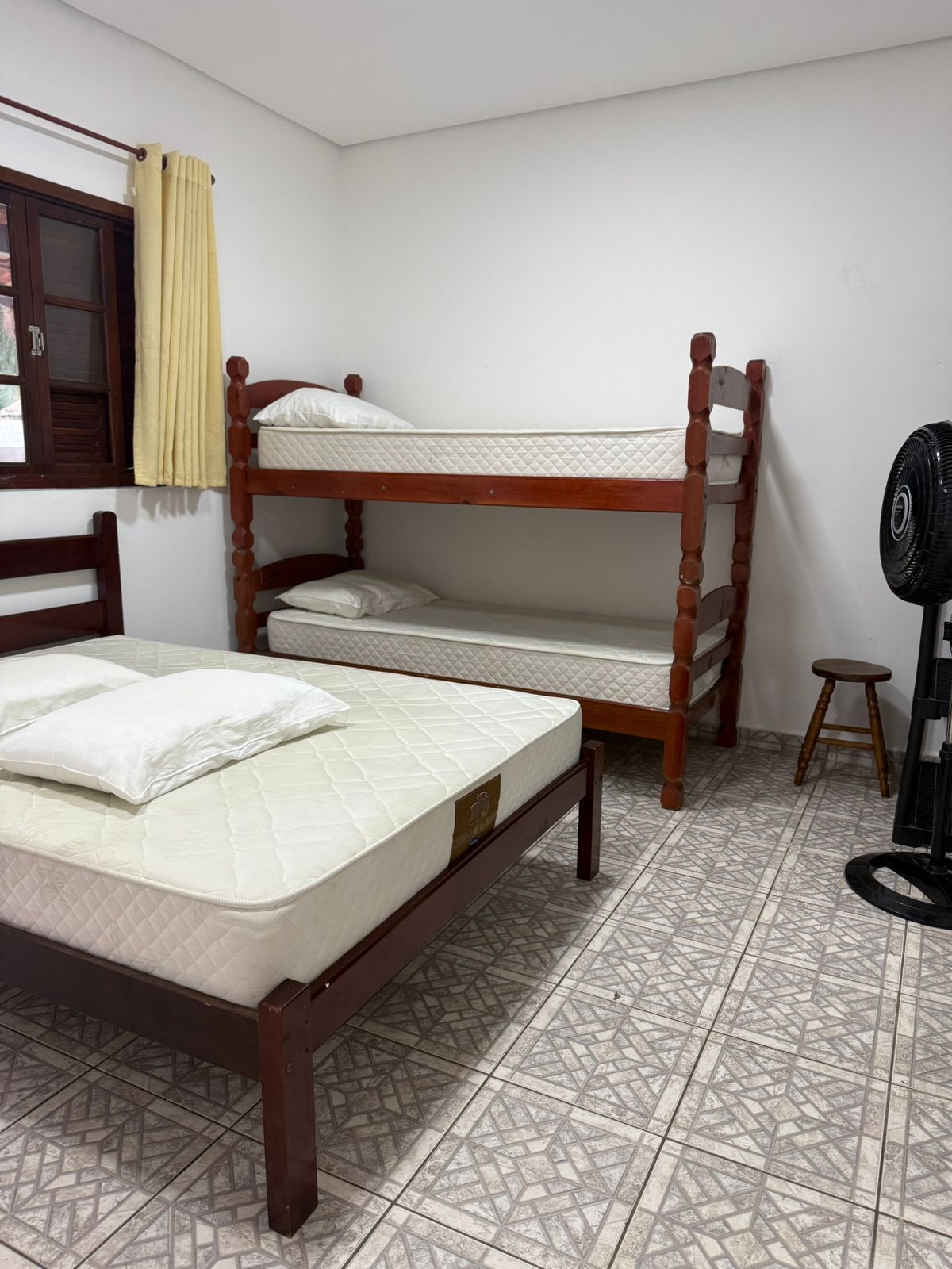 Quarto com beliches e uma cama de solteiro. Paredes claras, piso estampado e ventilador.
