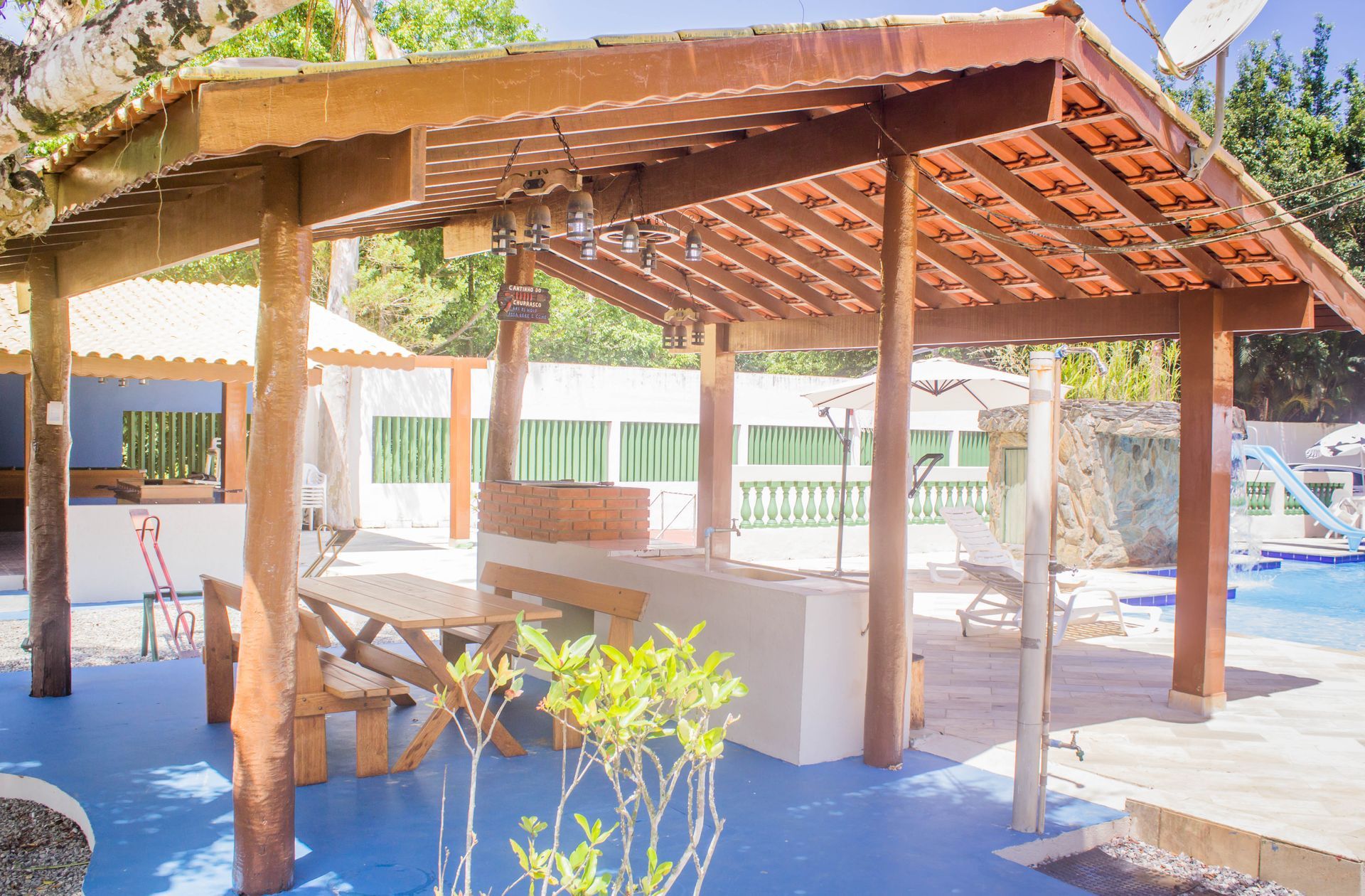 Gazebo de madeira com churrasqueira, mesa e assentos, ao lado de uma piscina. Ambiente externo ensolarado.