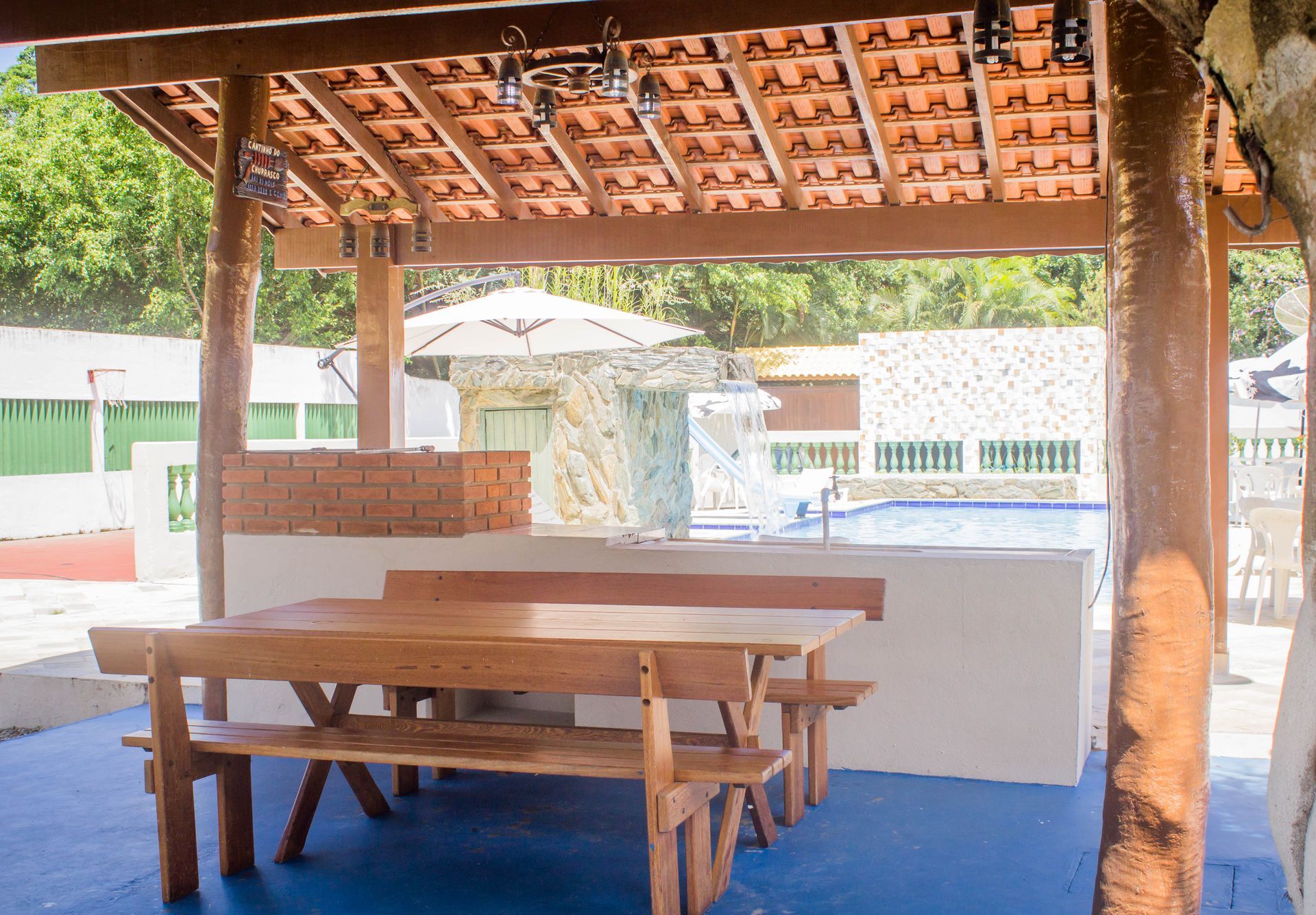 Área de jantar externa coberta com mesa de madeira, bancos e vista para a piscina.