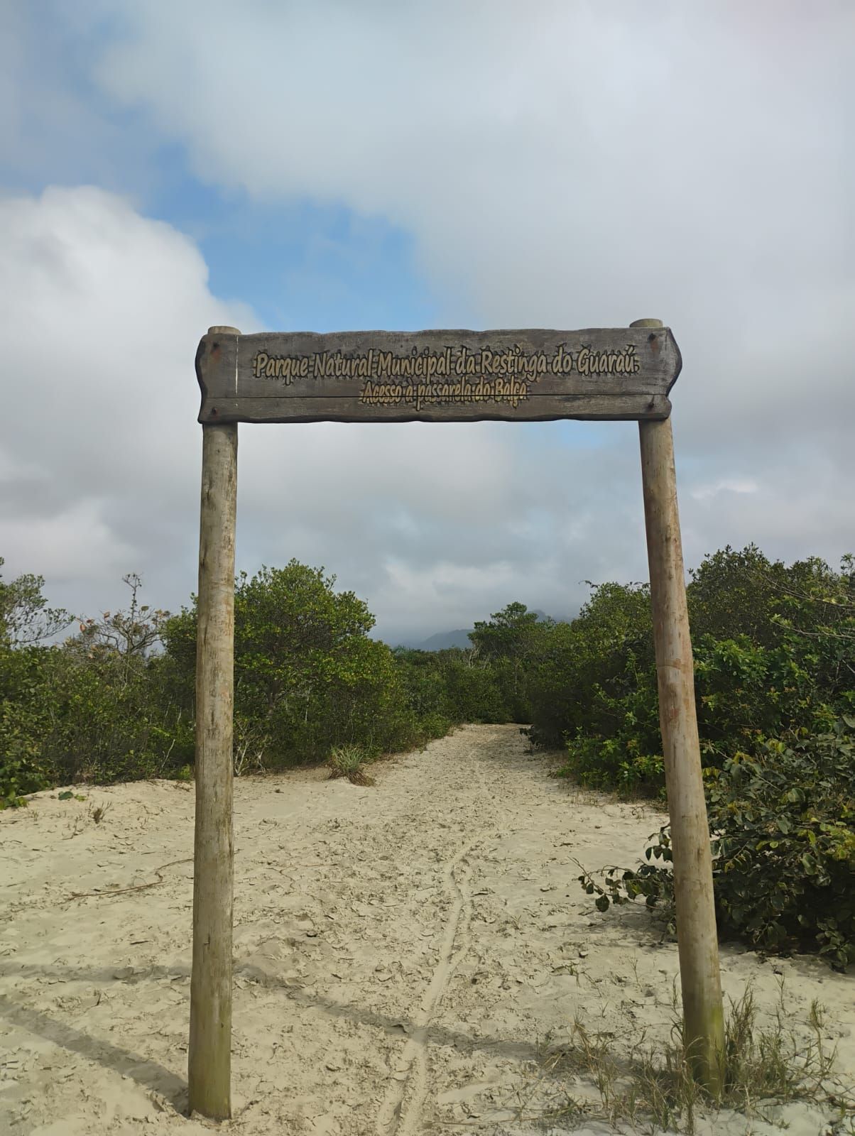 Placa de entrada de madeira para um parque nacional, com um caminho de areia e folhagem verde sob um céu nublado.