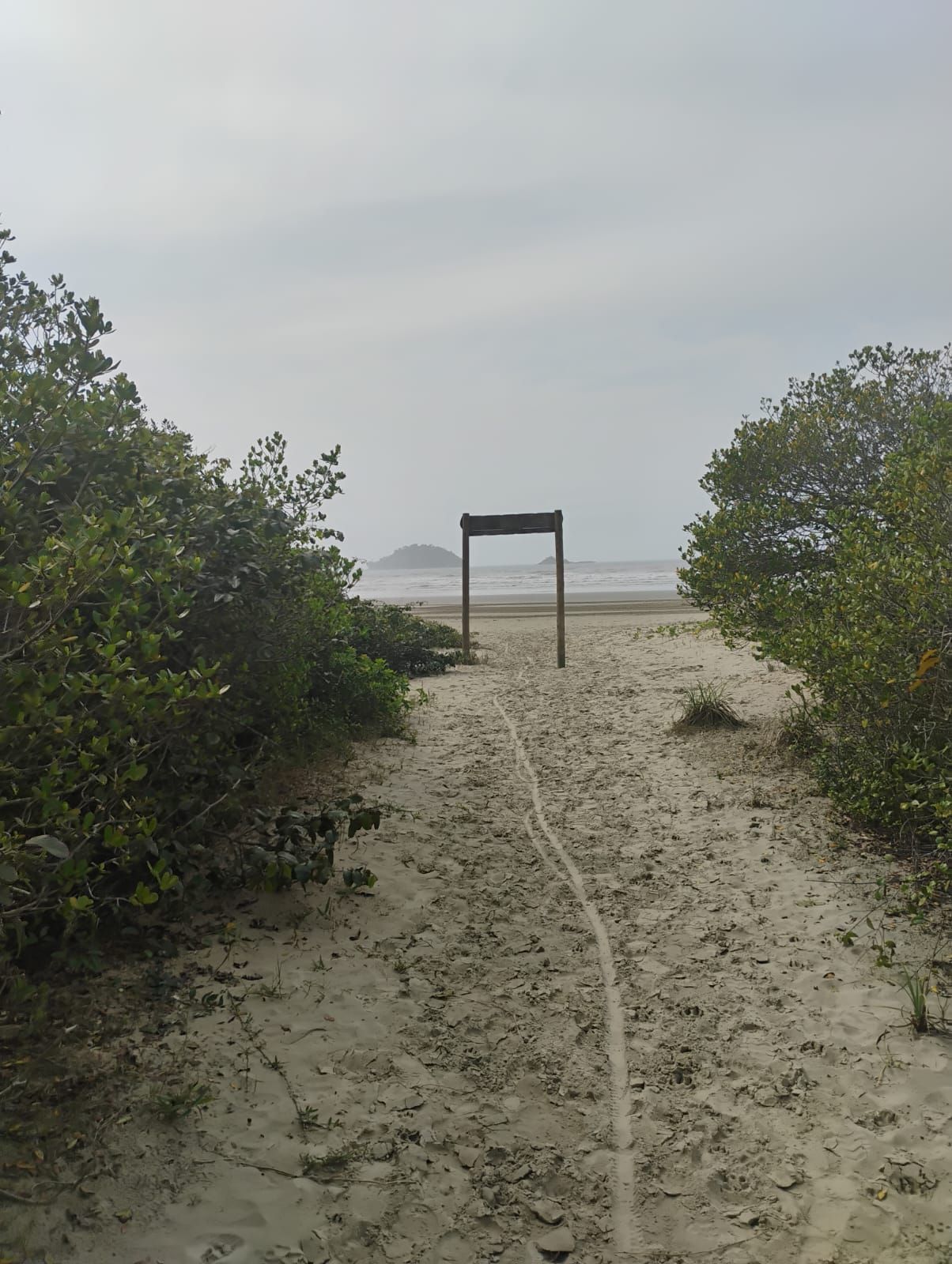 Caminho de areia que leva a uma praia e a um arco de madeira sob um céu nublado. Arbustos verdejantes margeiam o caminho.