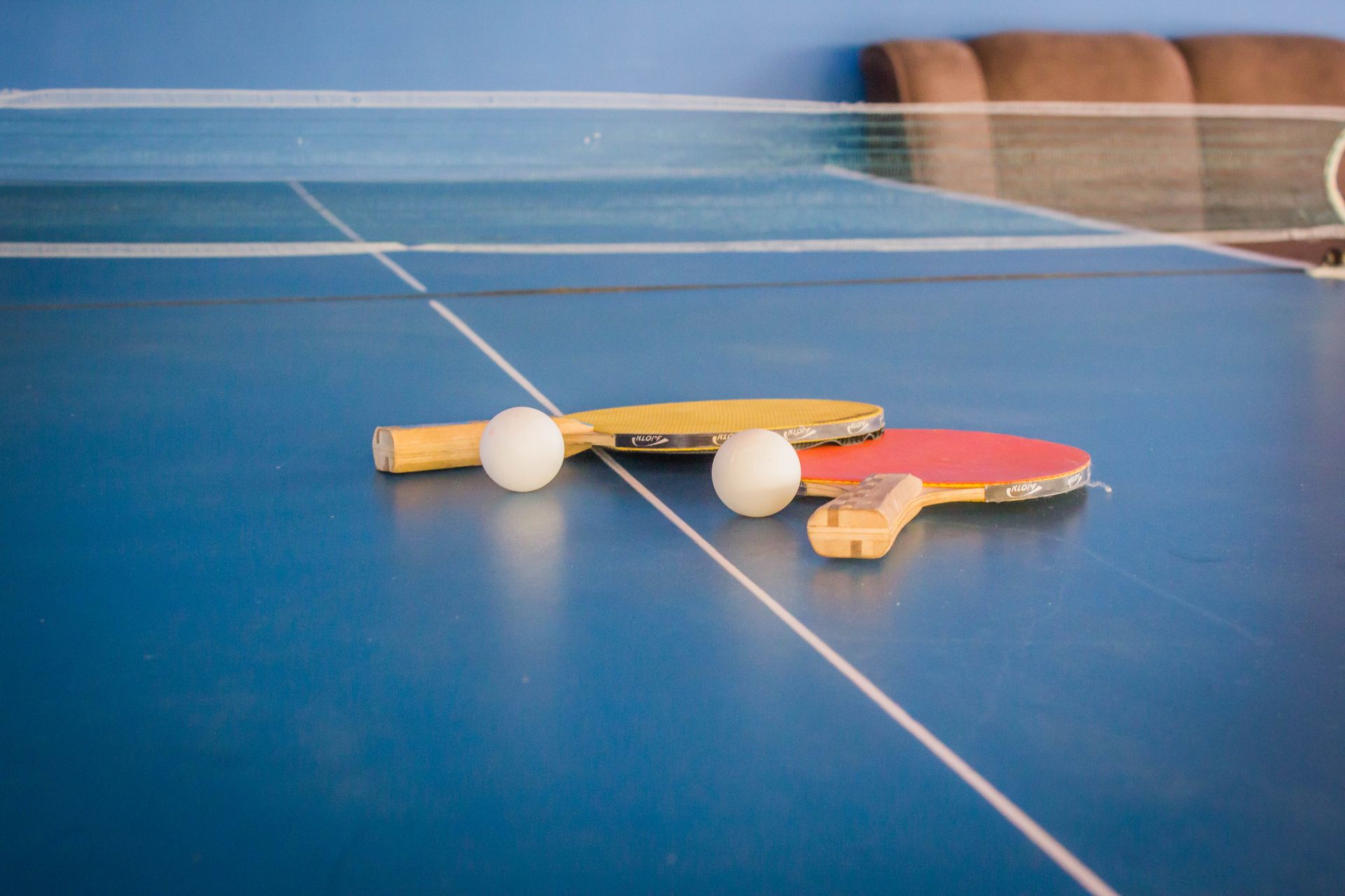 Mesa de pingue-pongue com duas raquetes e duas bolas brancas, em ambiente interno.