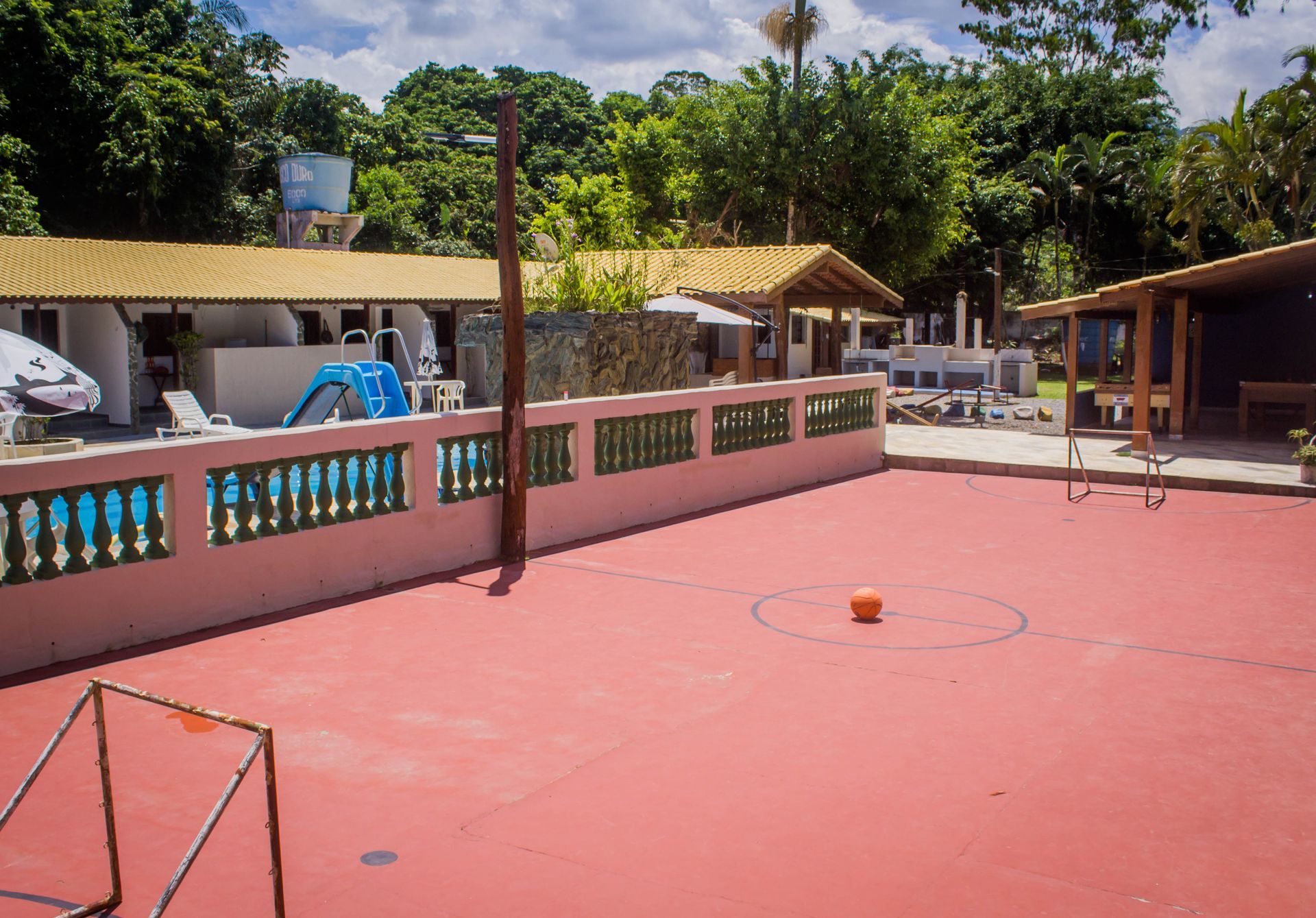 Quadra externa vermelha com uma bola, um gol e prédios ao fundo. Parede rosa com detalhes em azul.